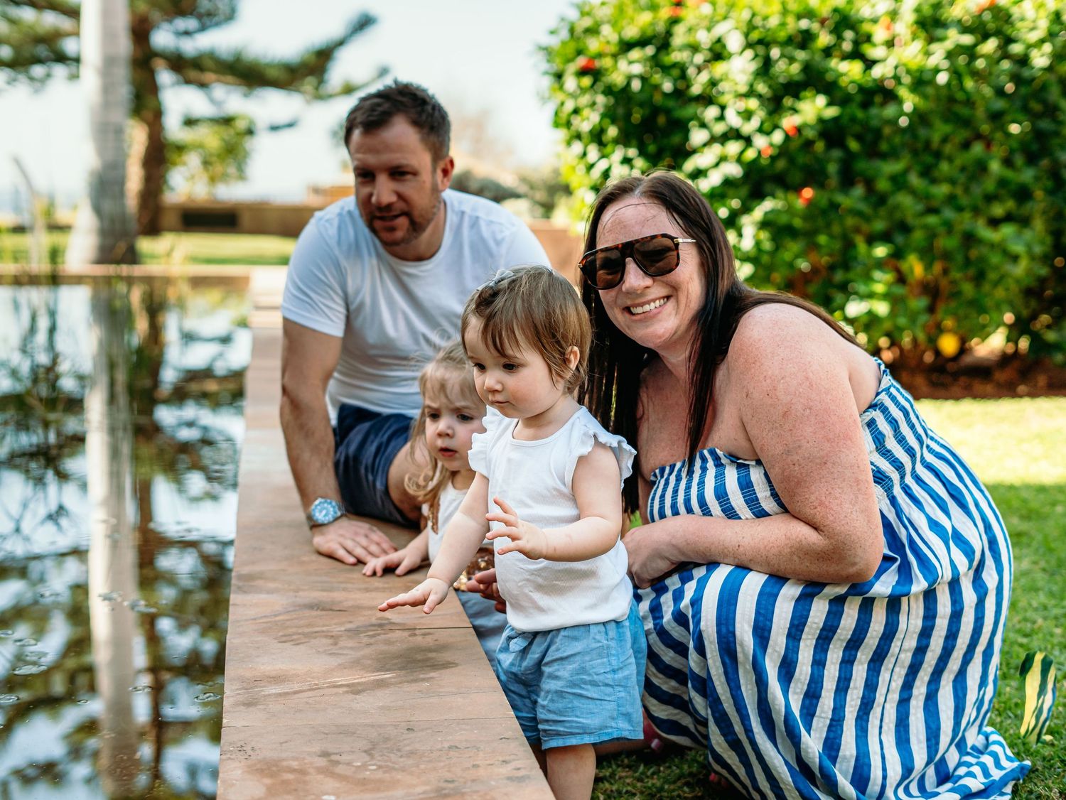 Smiling family of four posing together by a wooden ledge beside a peaceful garden pond.