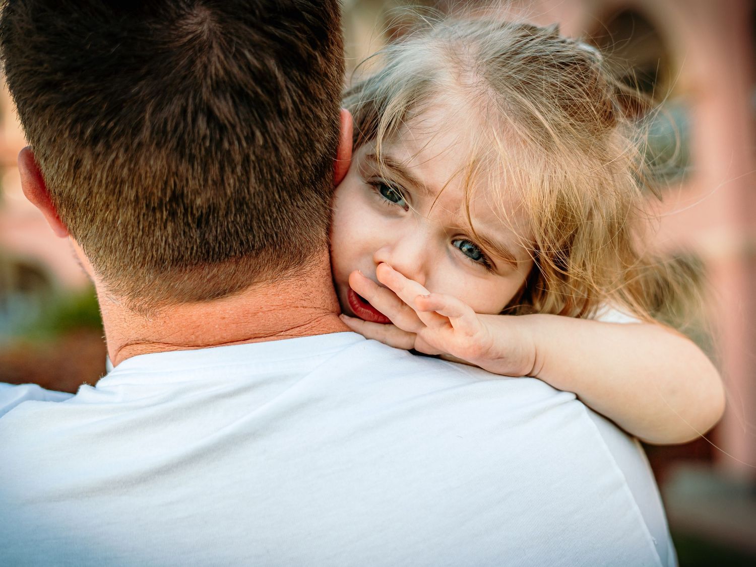 Emotional blonde toddler girl with blue eyes resting on fathers shoulder looking sad.
