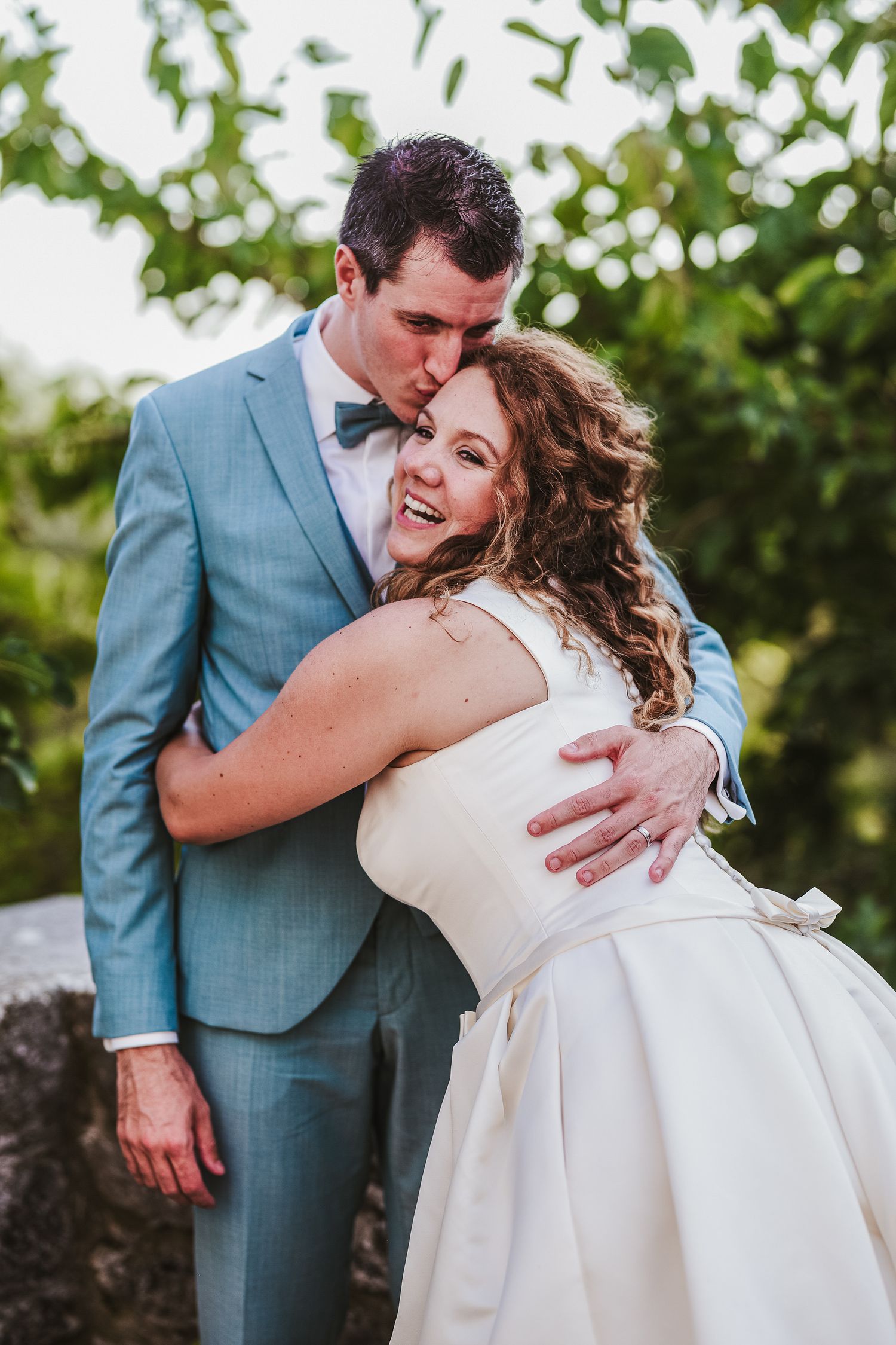 Groom in light blue suit kissing bride in white dress as she laughs joyfully outdoors.