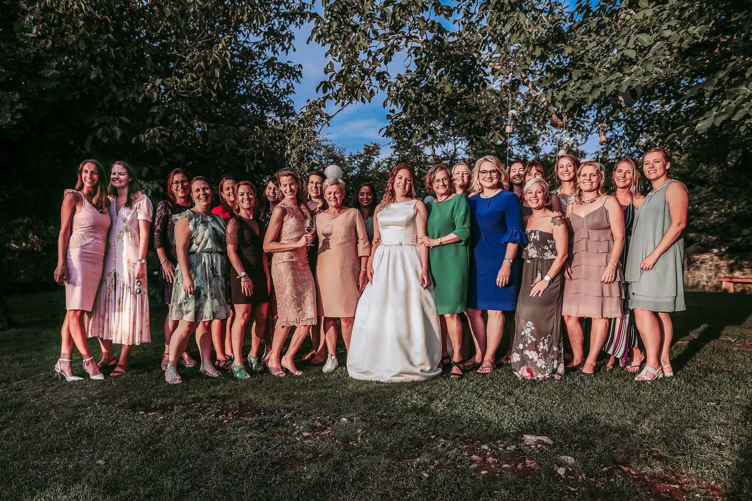 Bride in white gown posing with a large group of female wedding guests outdoors under trees.