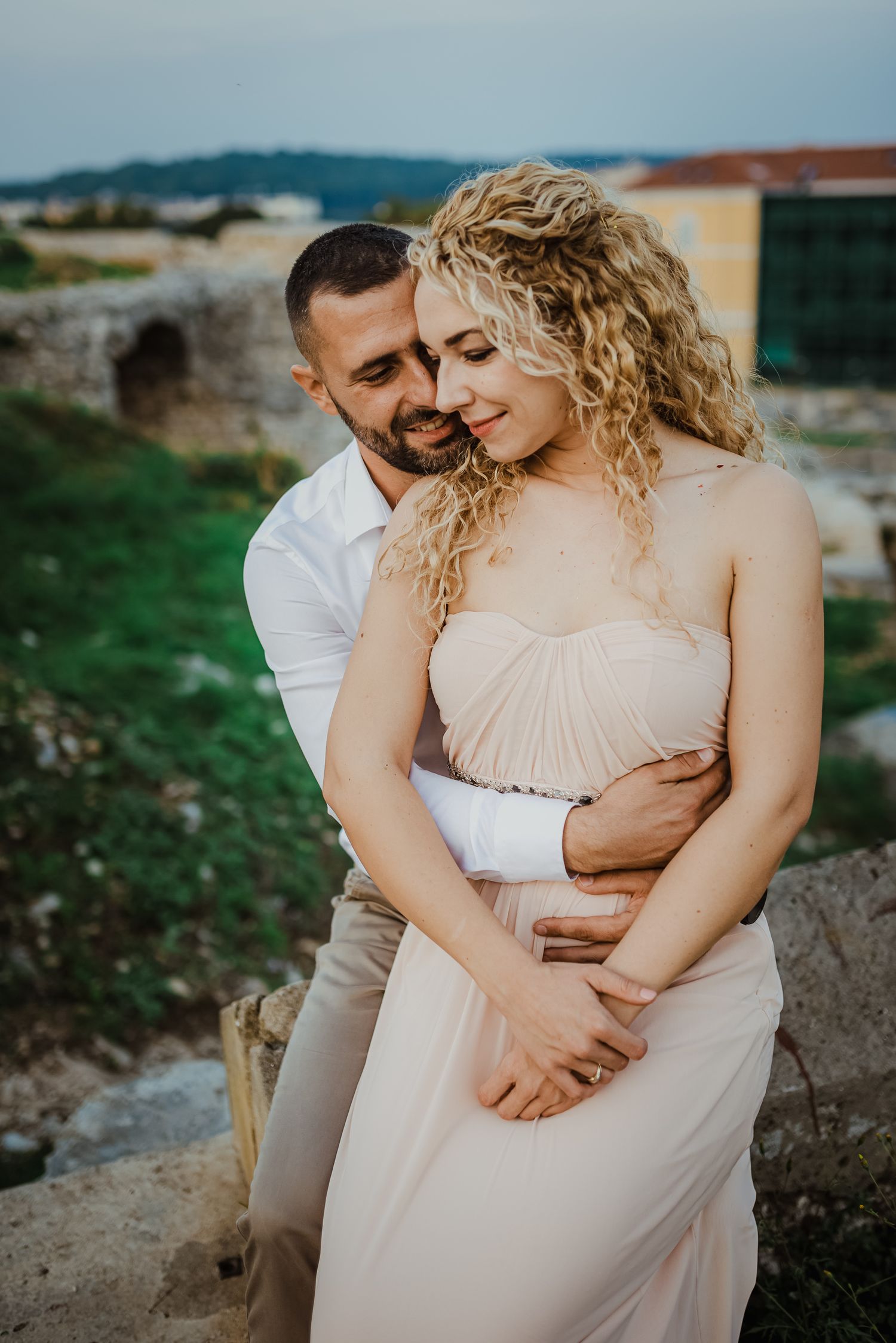 Groom embracing bride in strapless blush gown with curly hair at romantic outdoor wedding venue.