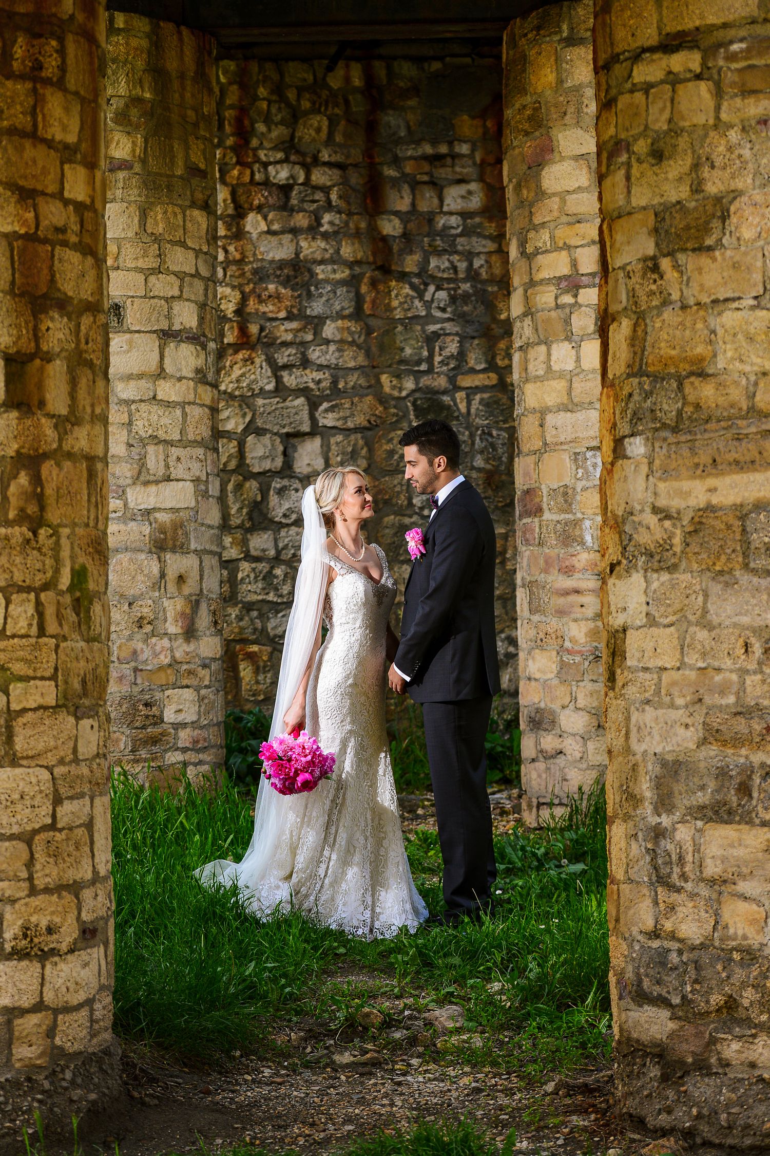 Wedding couple holding pink bouquet standing between rustic brick columns in romantic embrace.