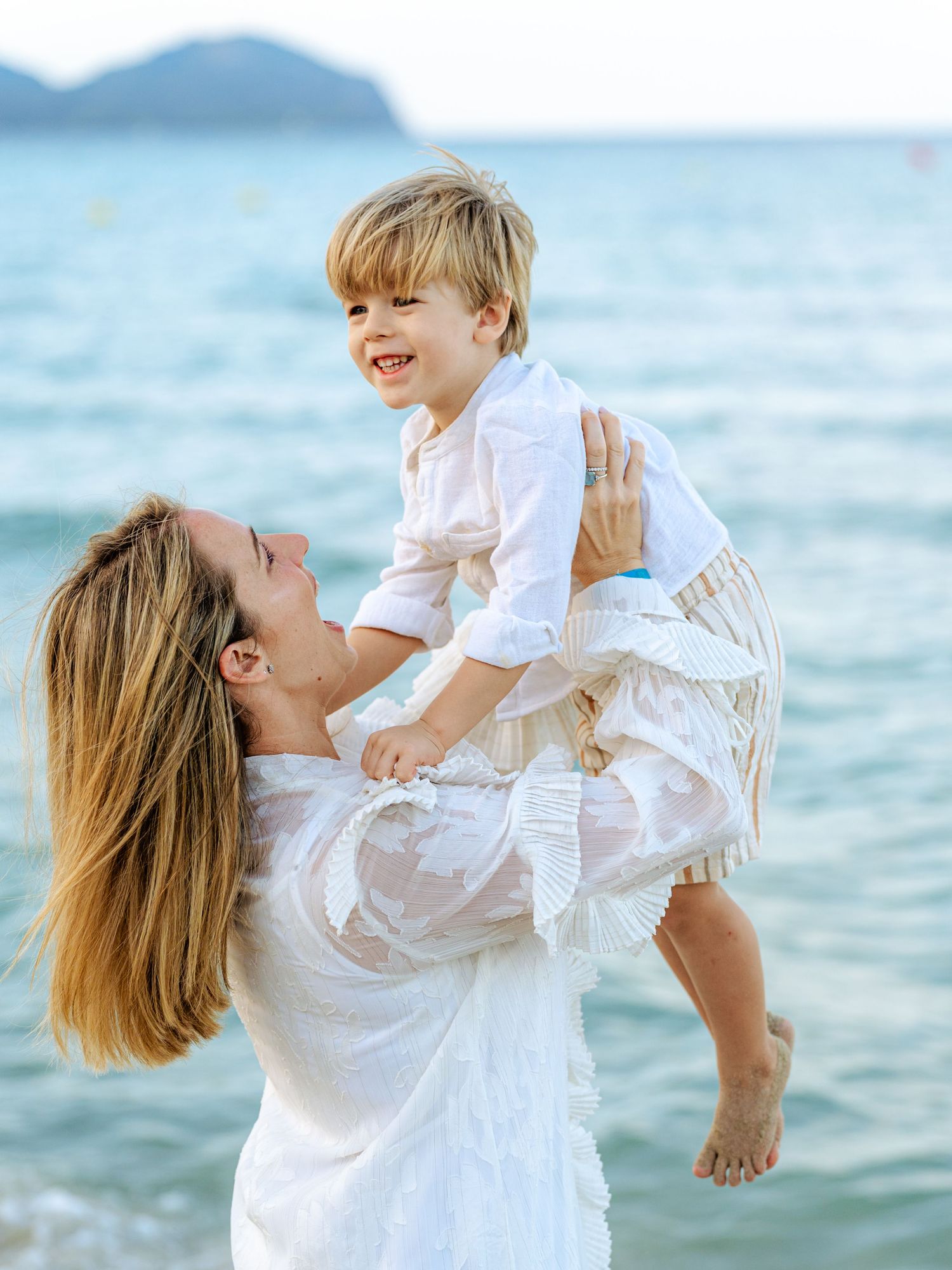 Smiling mother in white dress holding up happy toddler boy at seaside.