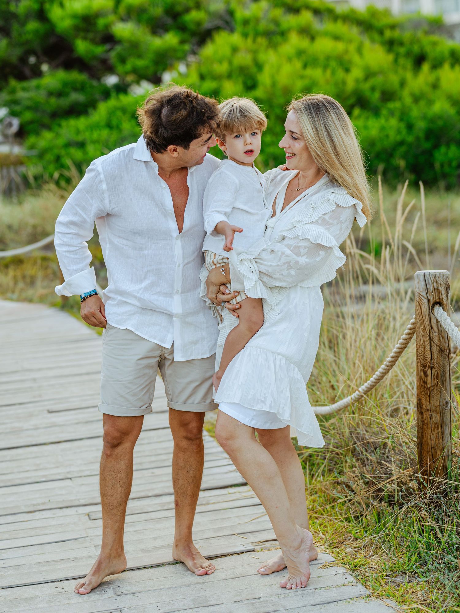 Happy family of three in white outfits on wooden boardwalk, parents holding young child.