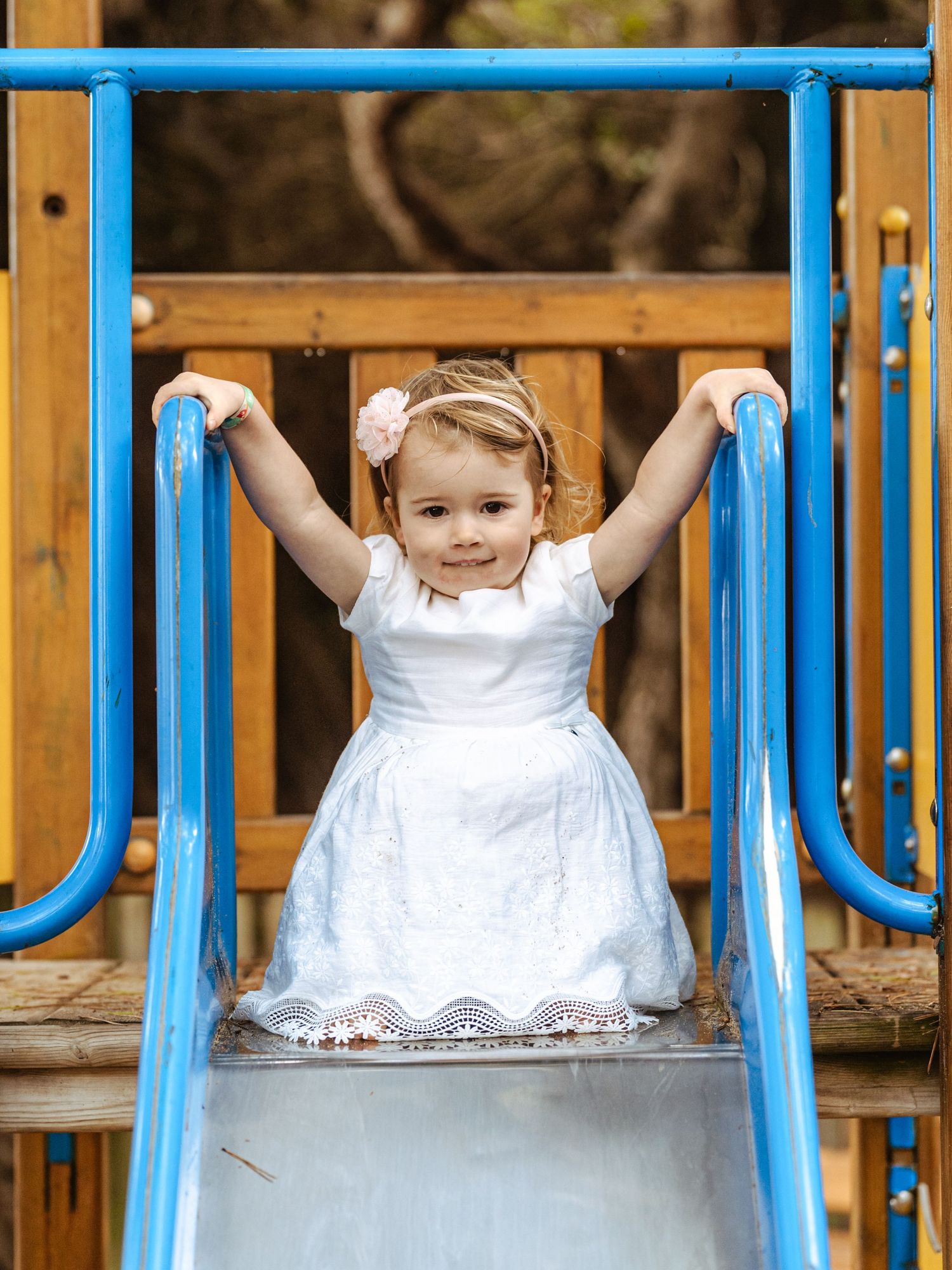 Little girl in white dress smiling on playground slide.