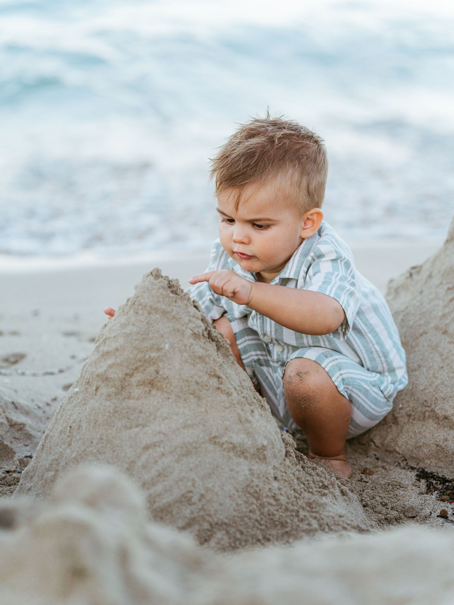Young toddler boy in striped romper plays and explores a sandcastle on the beach.