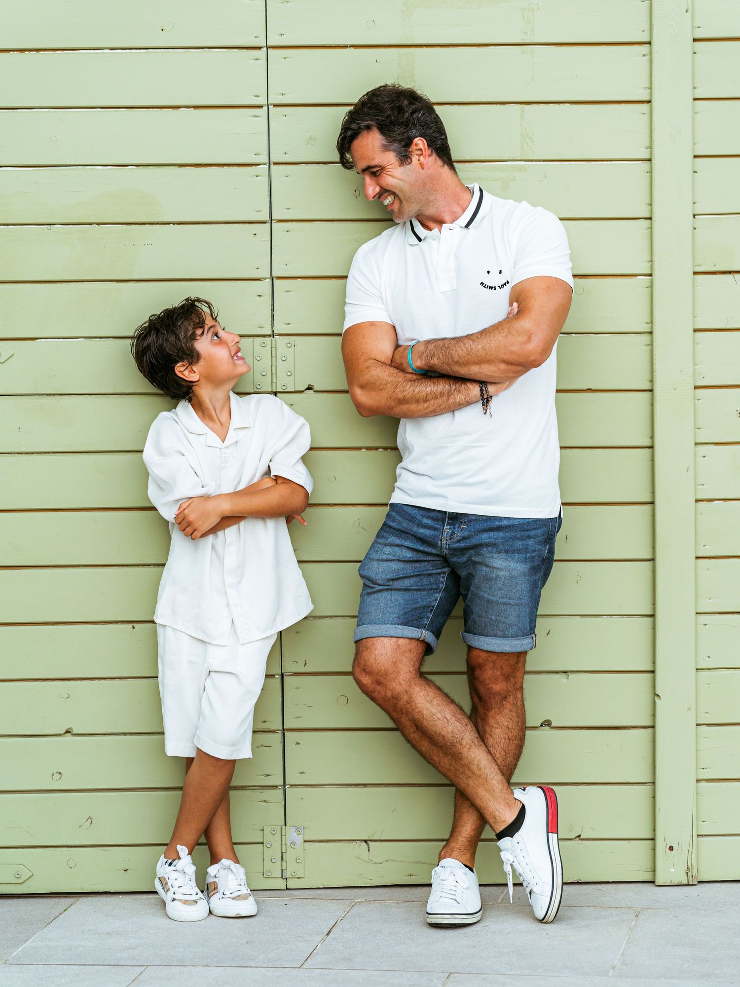 Father and son leaning against a green wall, smiling at each other in matching white outfits.