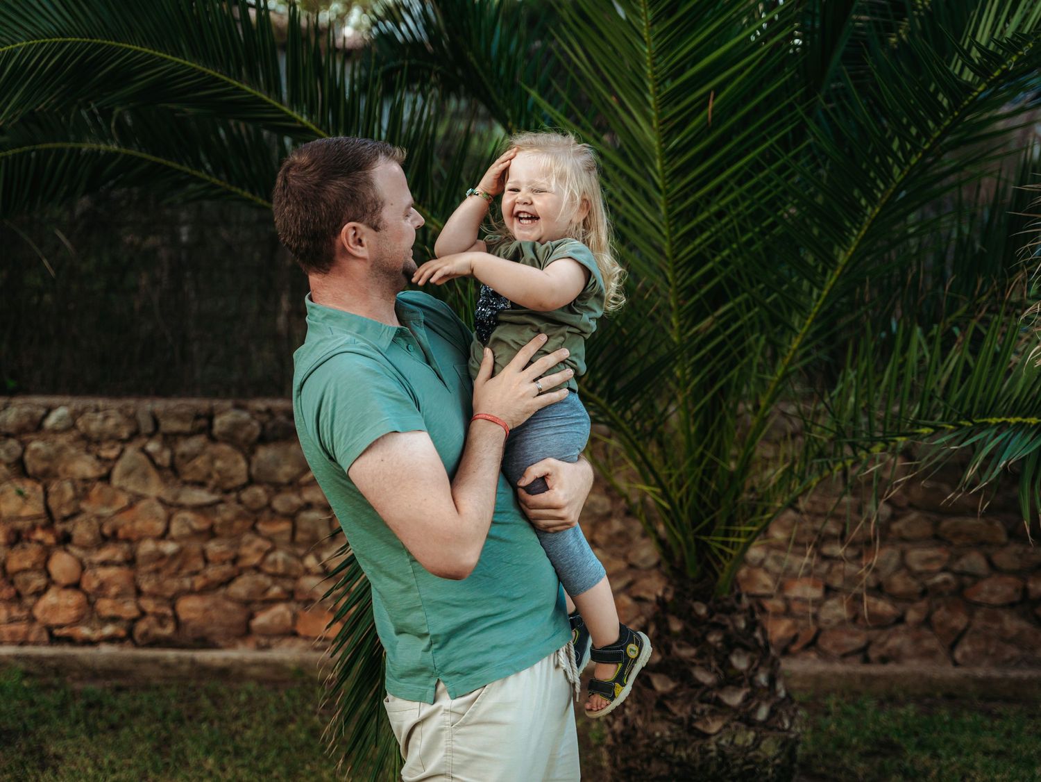 A laughing father in green shirt lifts his giggling toddler girl near a tropical palm tree.