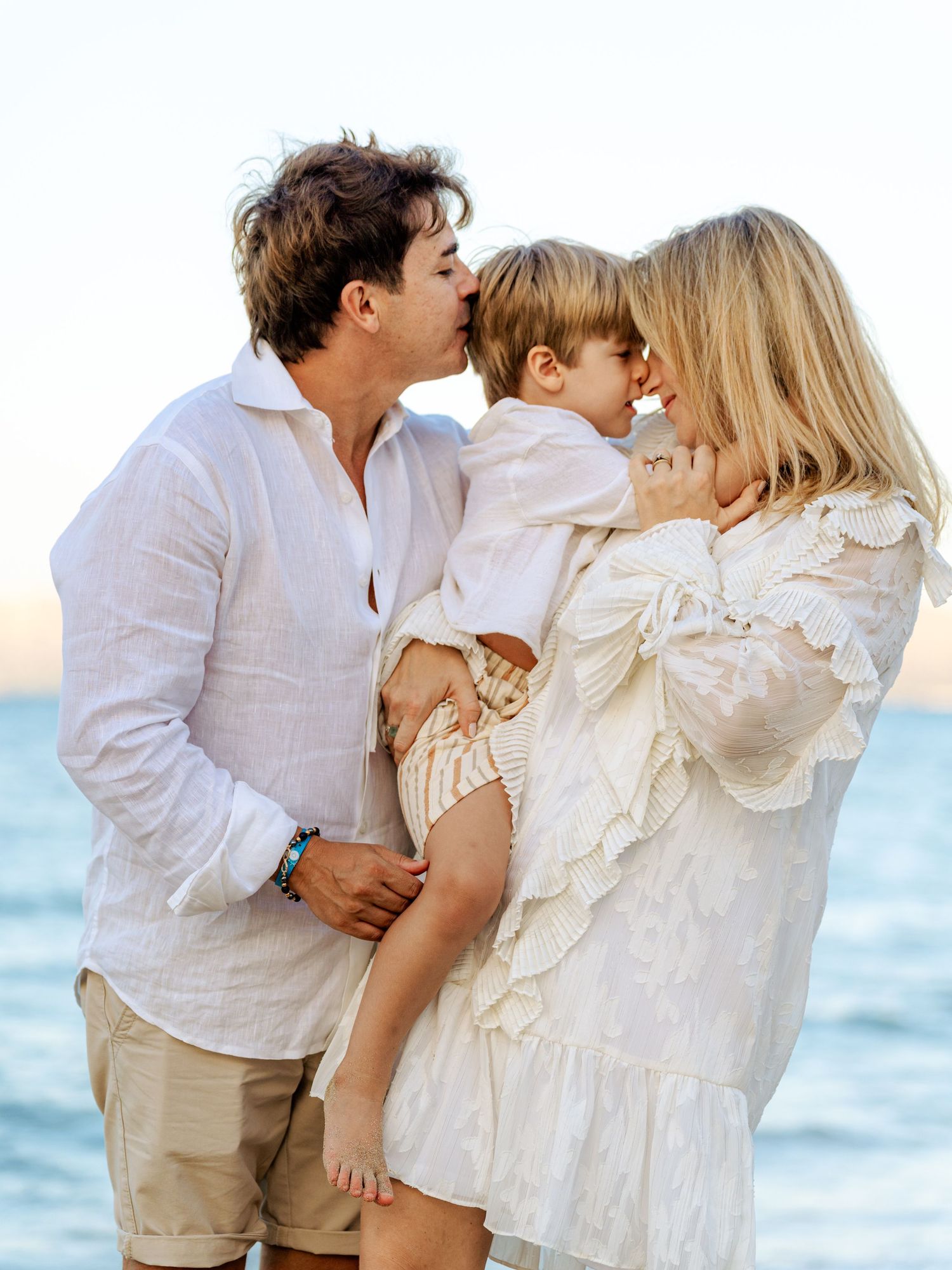 Parents kissing toddler child between them on scenic beach.