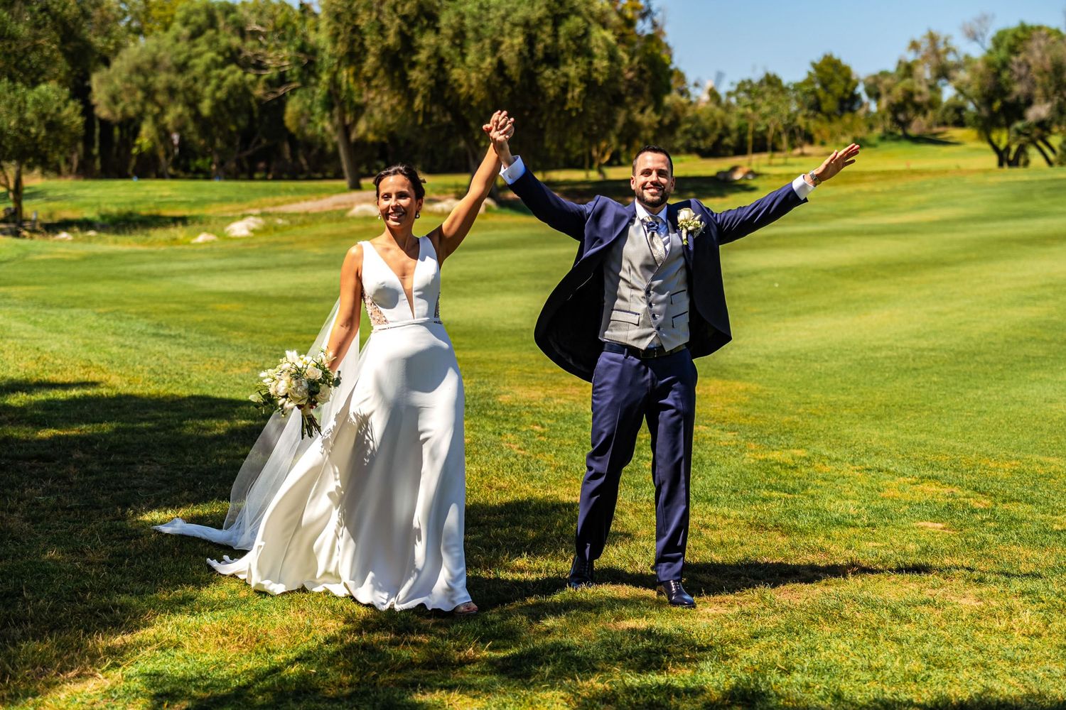 Joyful bride and groom celebrating with raised arms on a sunny green golf course.