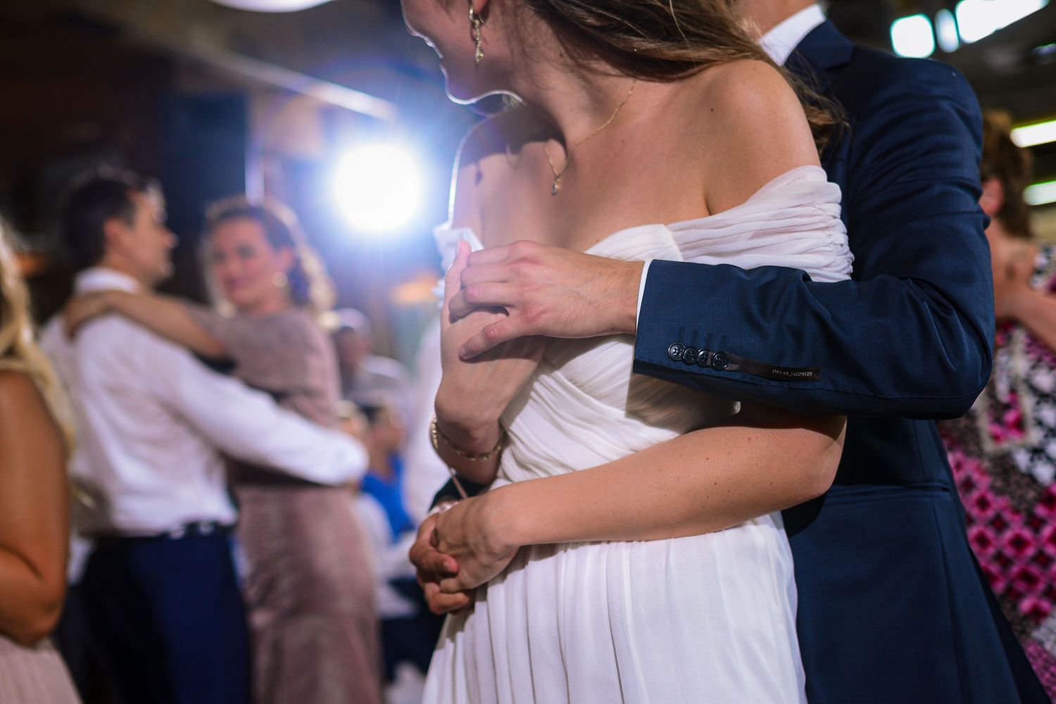 Groom in navy suit holds bride close during wedding reception first dance with guests in background.