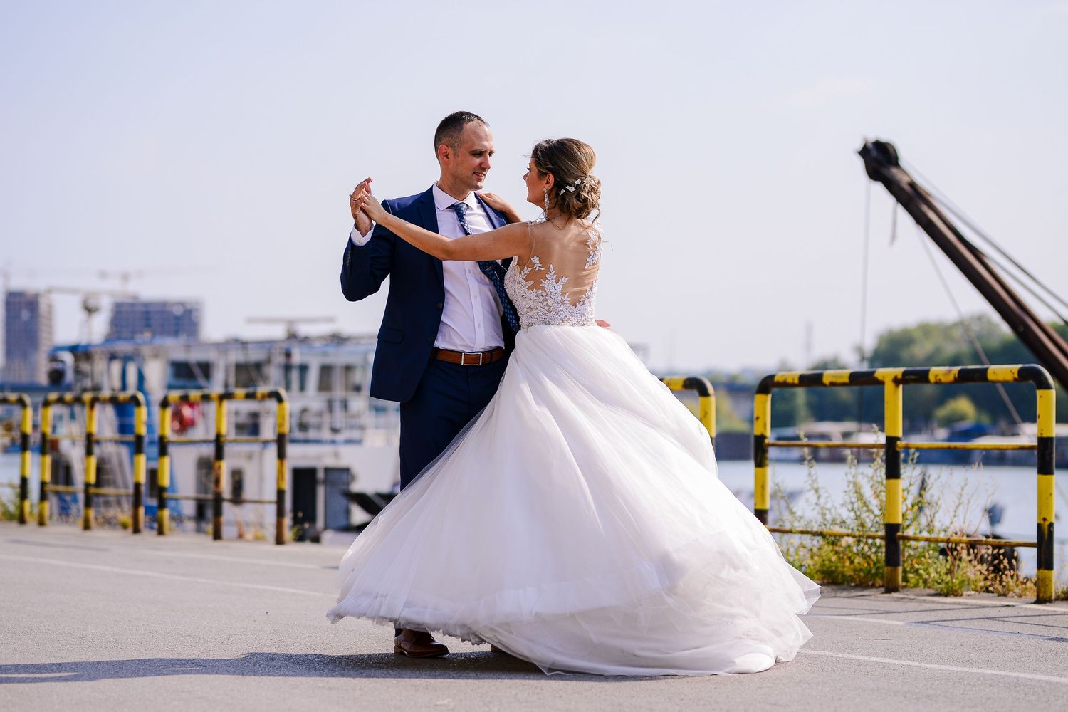 Bride and groom dancing outdoors near a sunny waterfront in wedding attire.