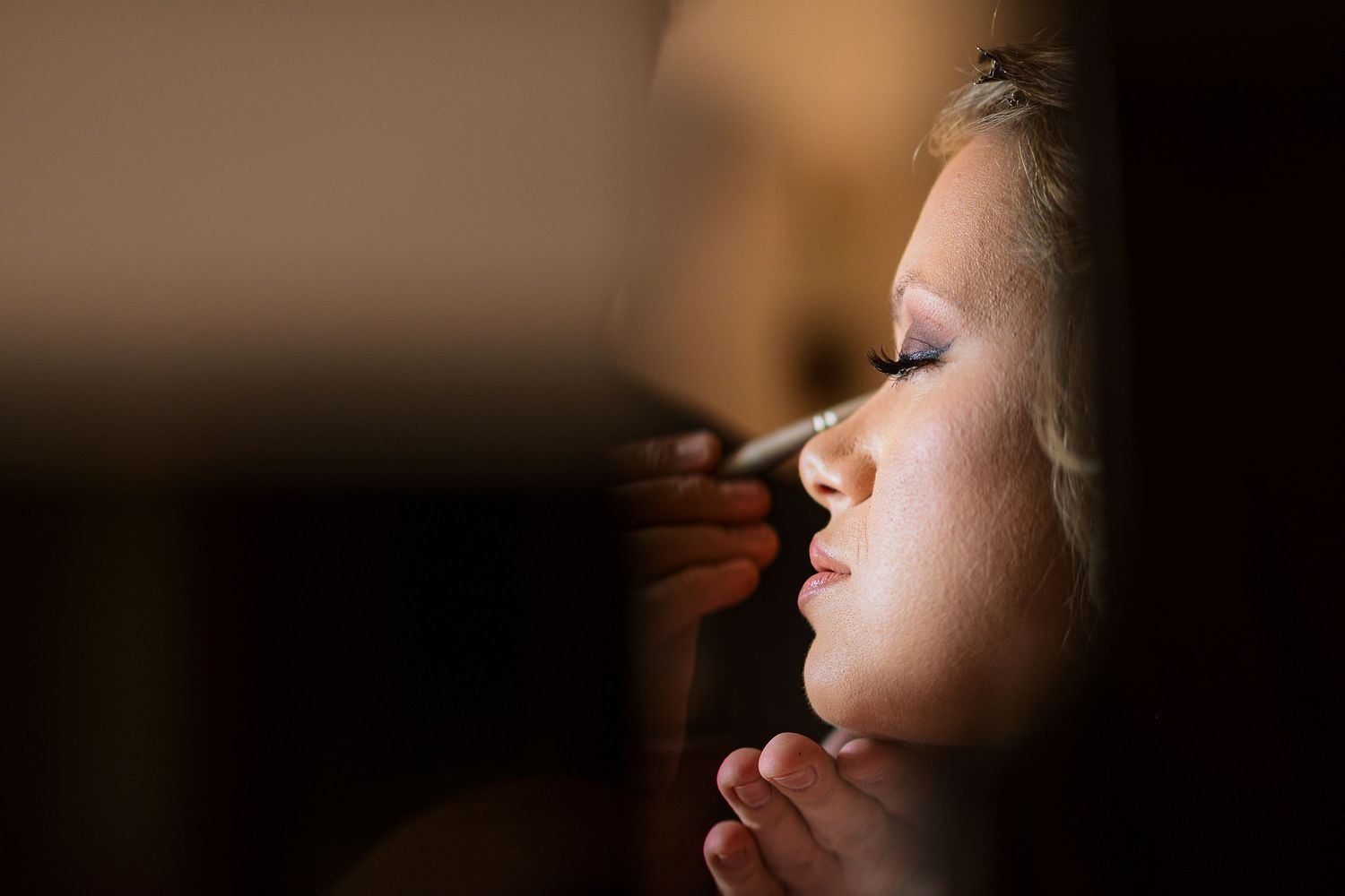 Close-up of a bride having makeup applied before her wedding ceremony in soft dramatic lighting.