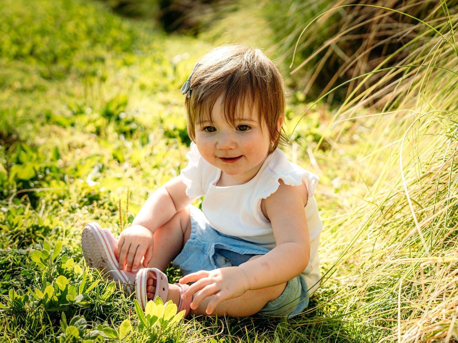 Adorable toddler girl sitting in green grass wearing white top and denim shorts.