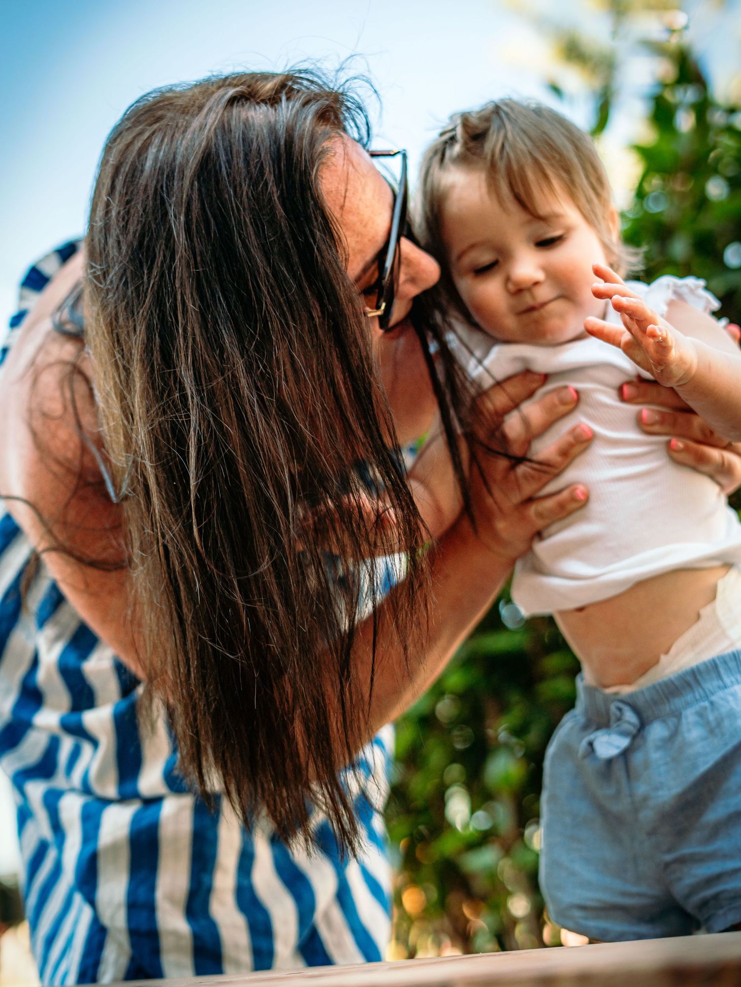 Loving mother holding and kissing her smiling baby outdoors in summer.