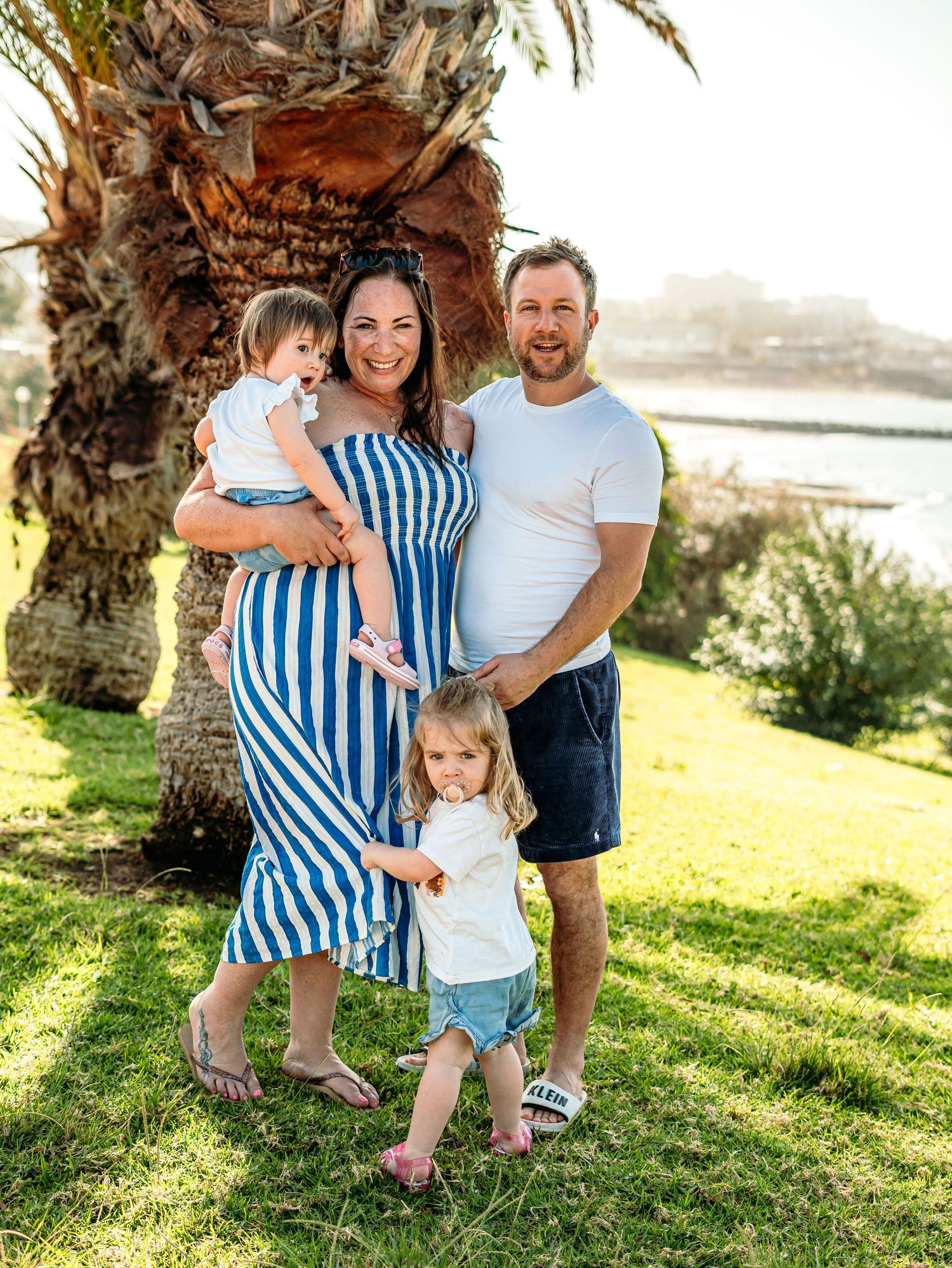 Happy family of four posing by a palm tree near the ocean on a sunny day.