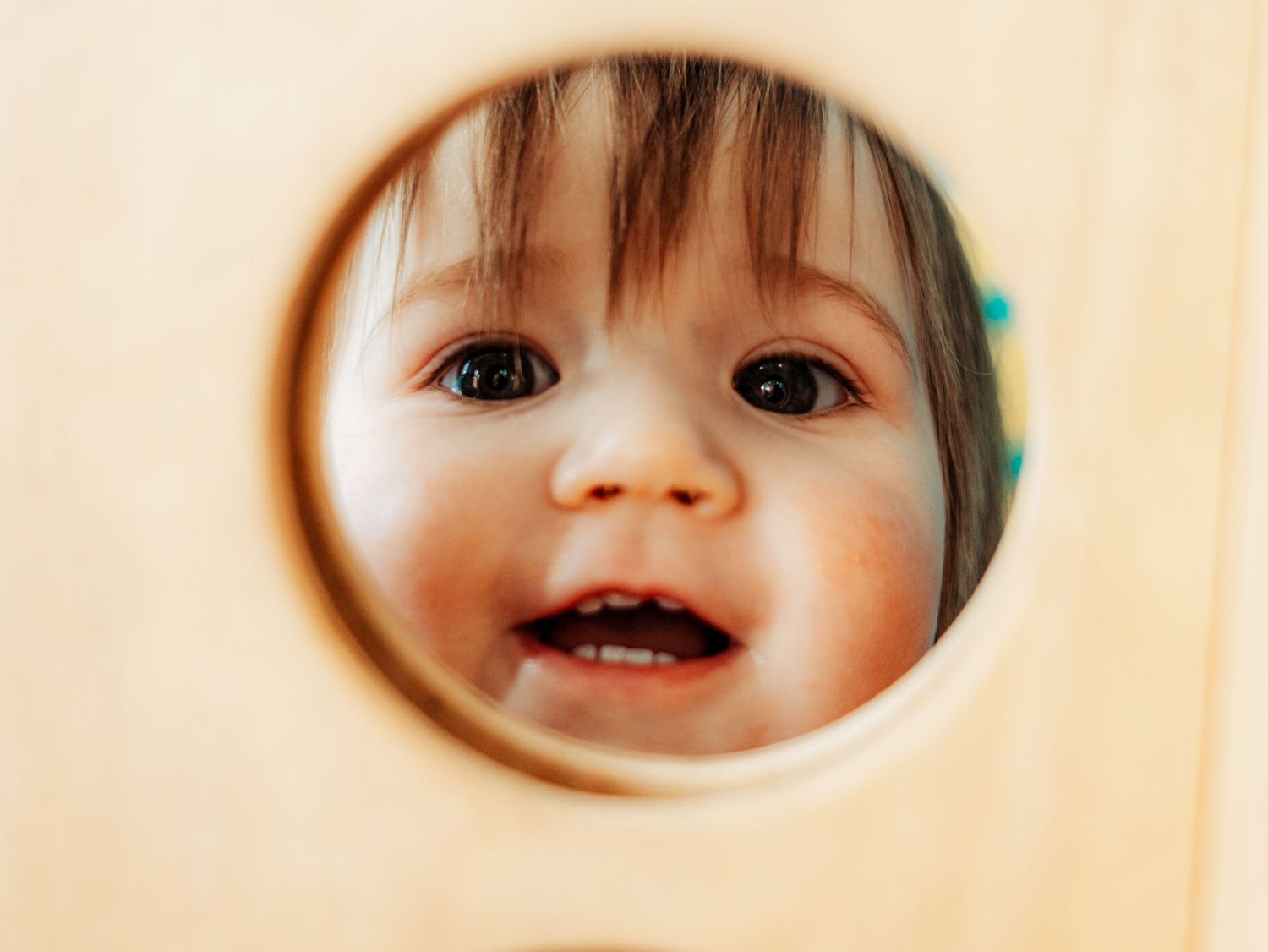 Smiling toddler peering through a circular hole in a wooden play structure showing tiny teeth.