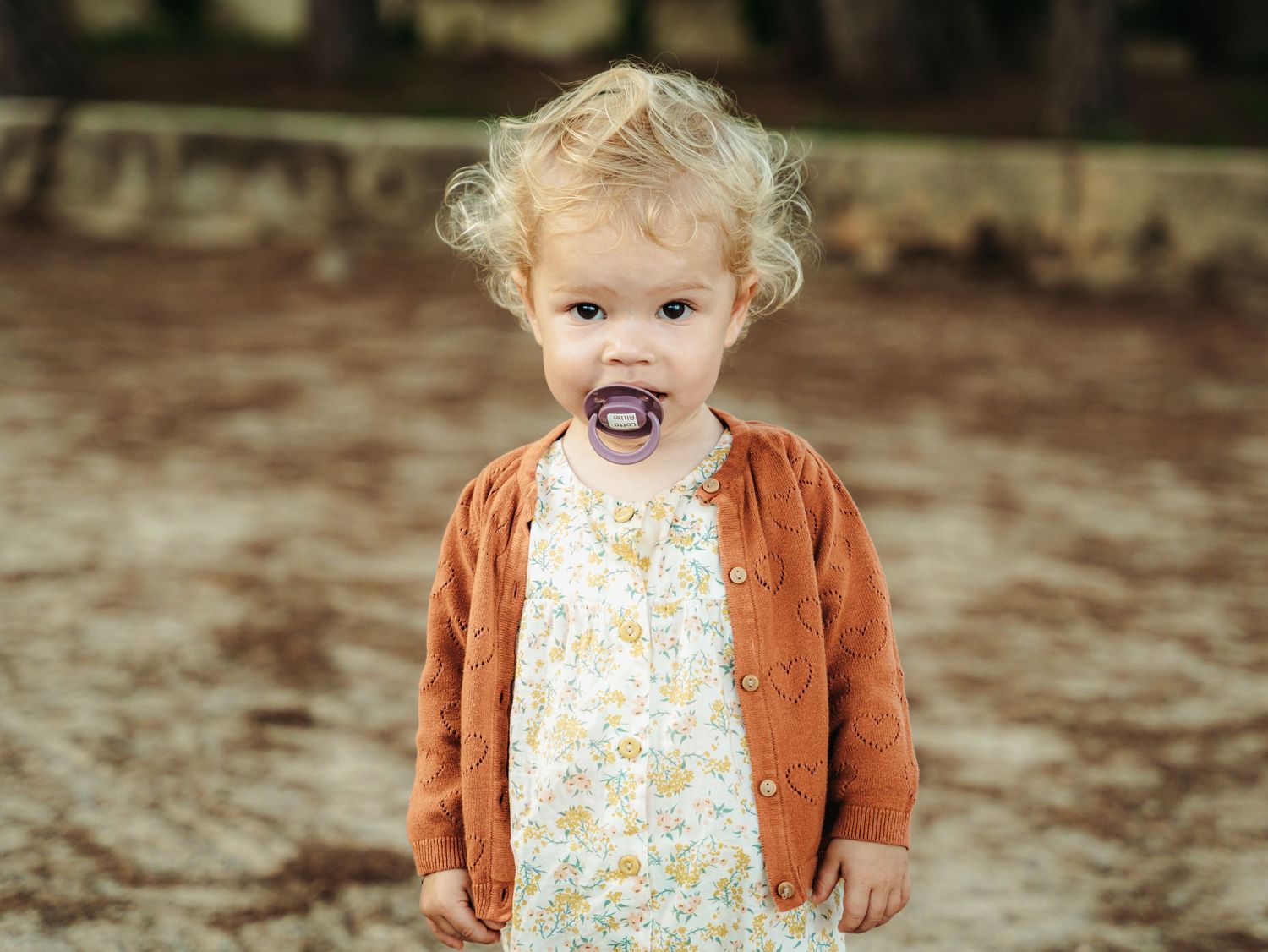 Close-up of curly-haired toddler in rust cardigan and floral dress with purple pacifier.