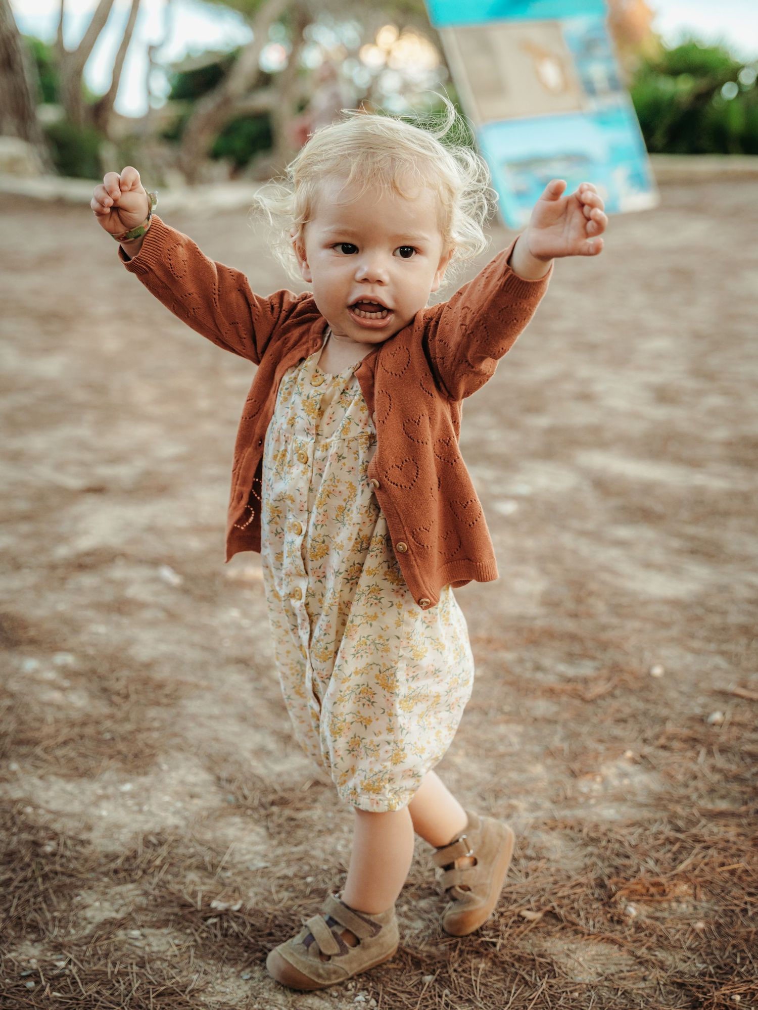 Adorable blonde toddler in floral romper and rust cardigan raising arms outdoors on sandy ground.