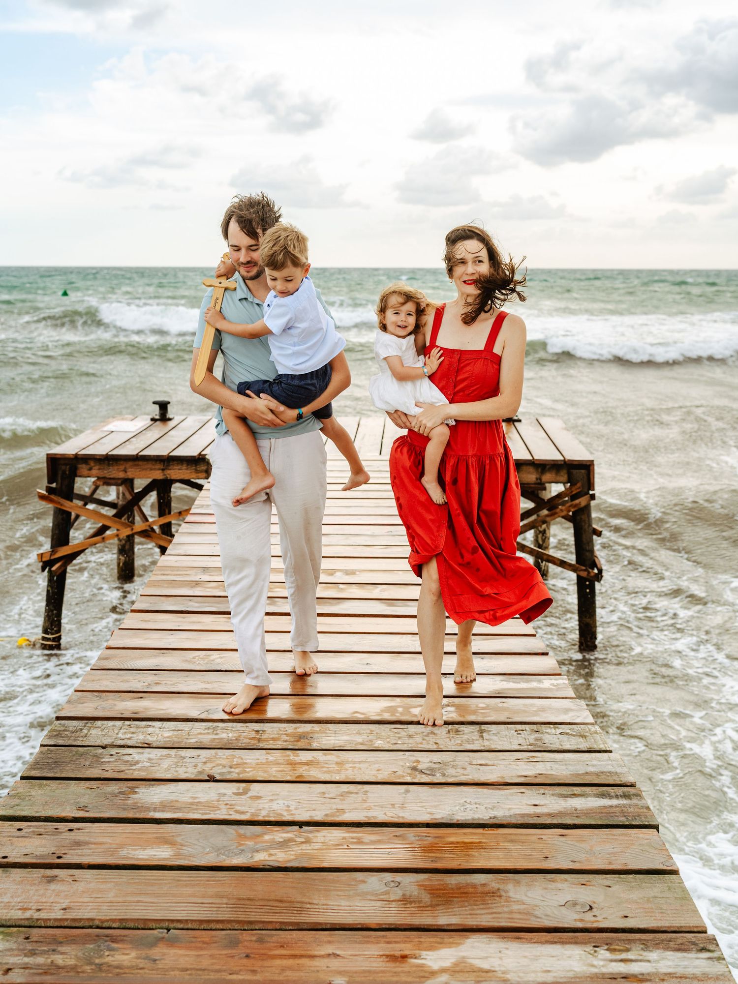Family of four on wooden beach pier, mom in red dress, dad holding son, waves in background.