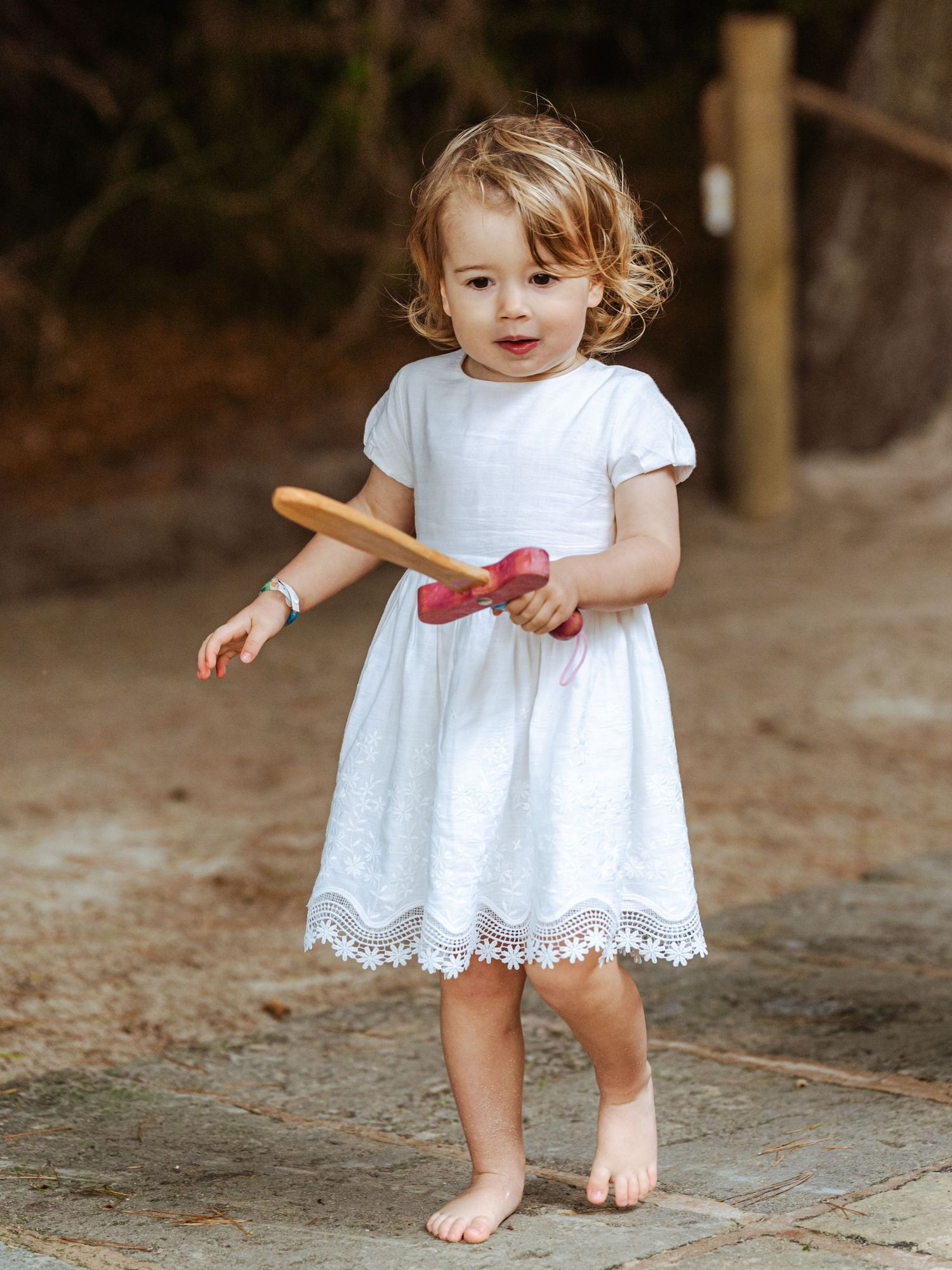 Cute toddler girl in white lace dress holding a wooden toy while standing barefoot outdoors.
