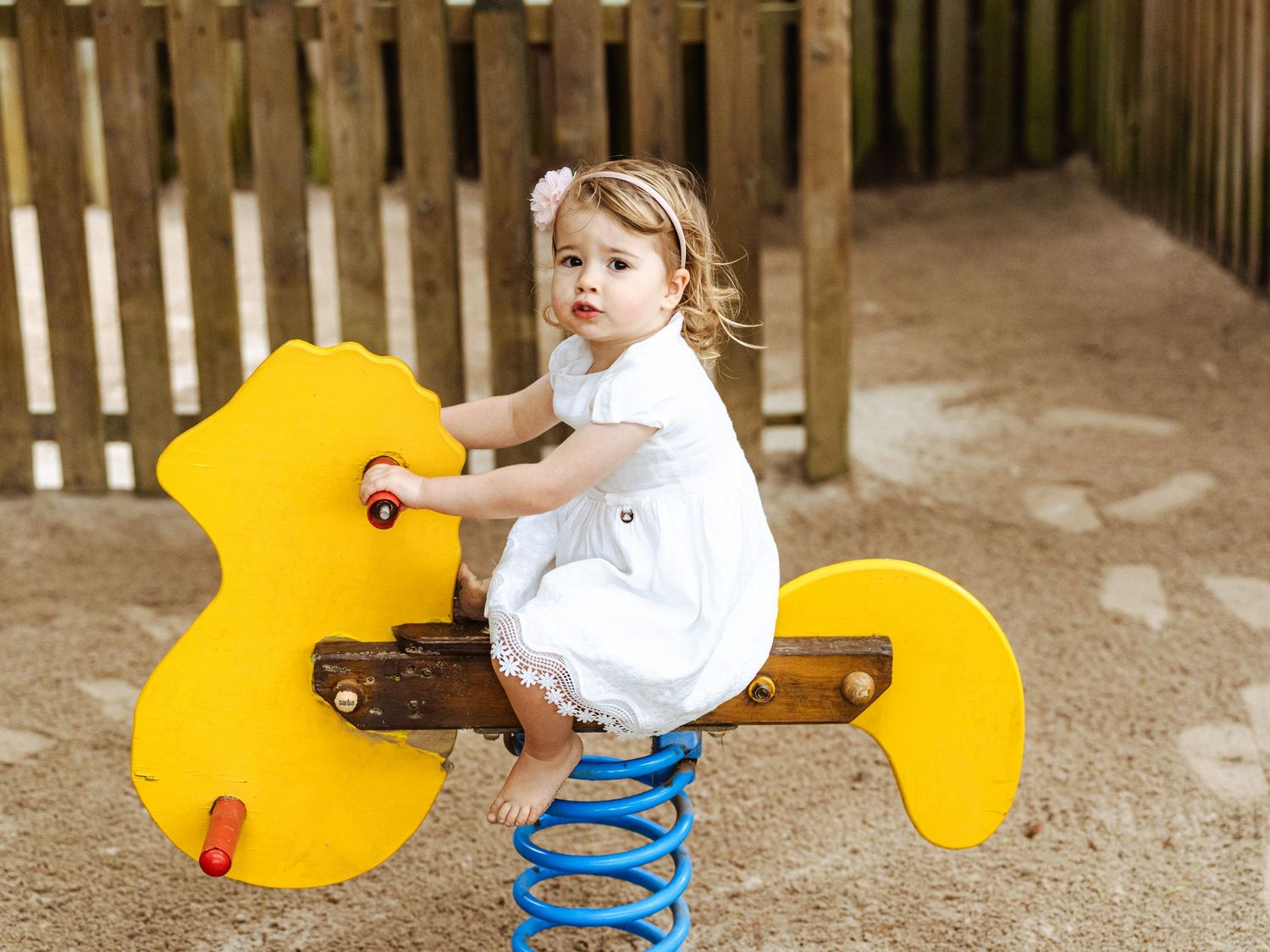 Adorable toddler girl in white dress riding yellow spring horse on sandy playground equipment outdoors.