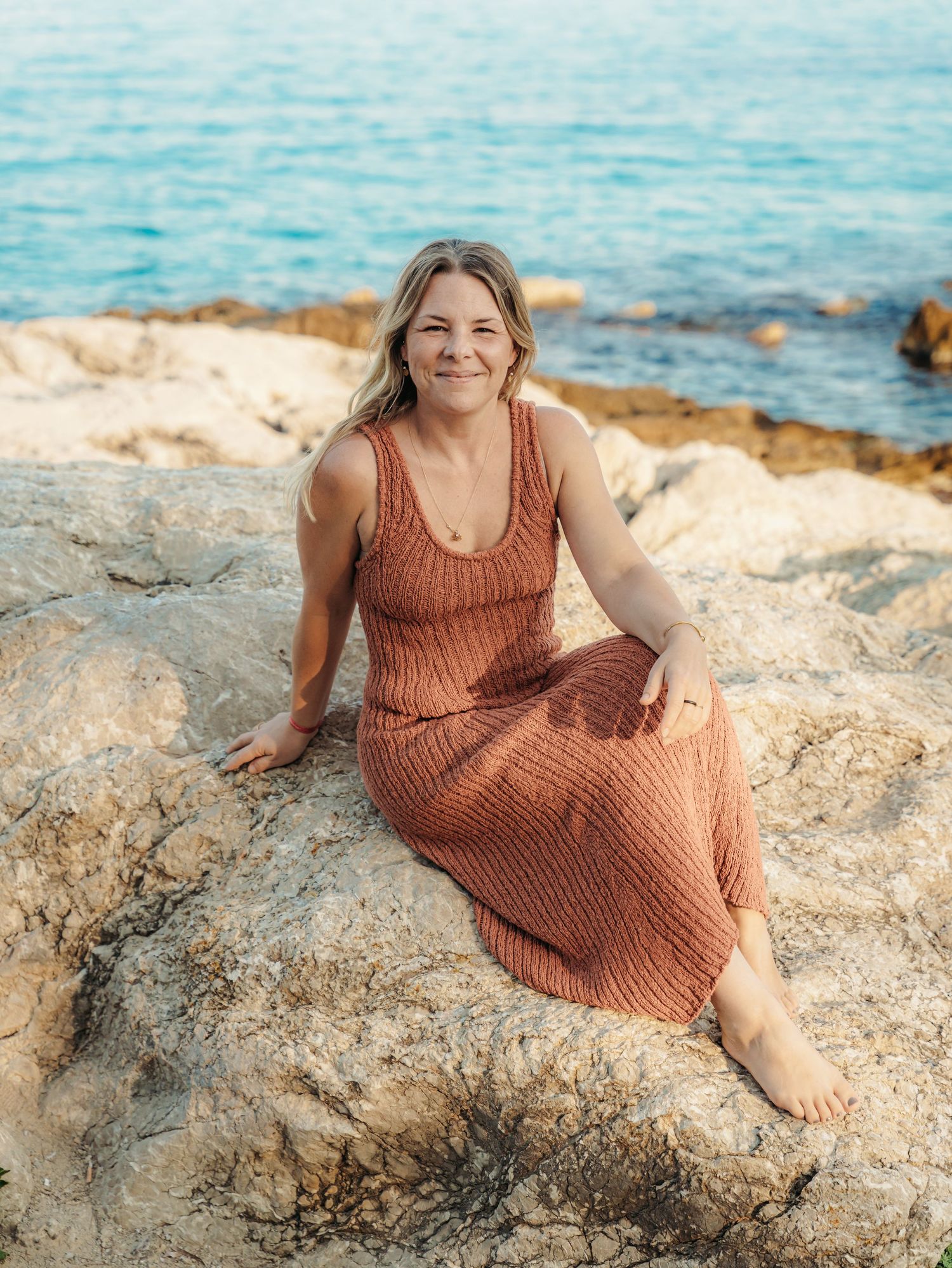 Smiling woman in terracotta knit dress sitting barefoot on sun-lit rocky coastline.