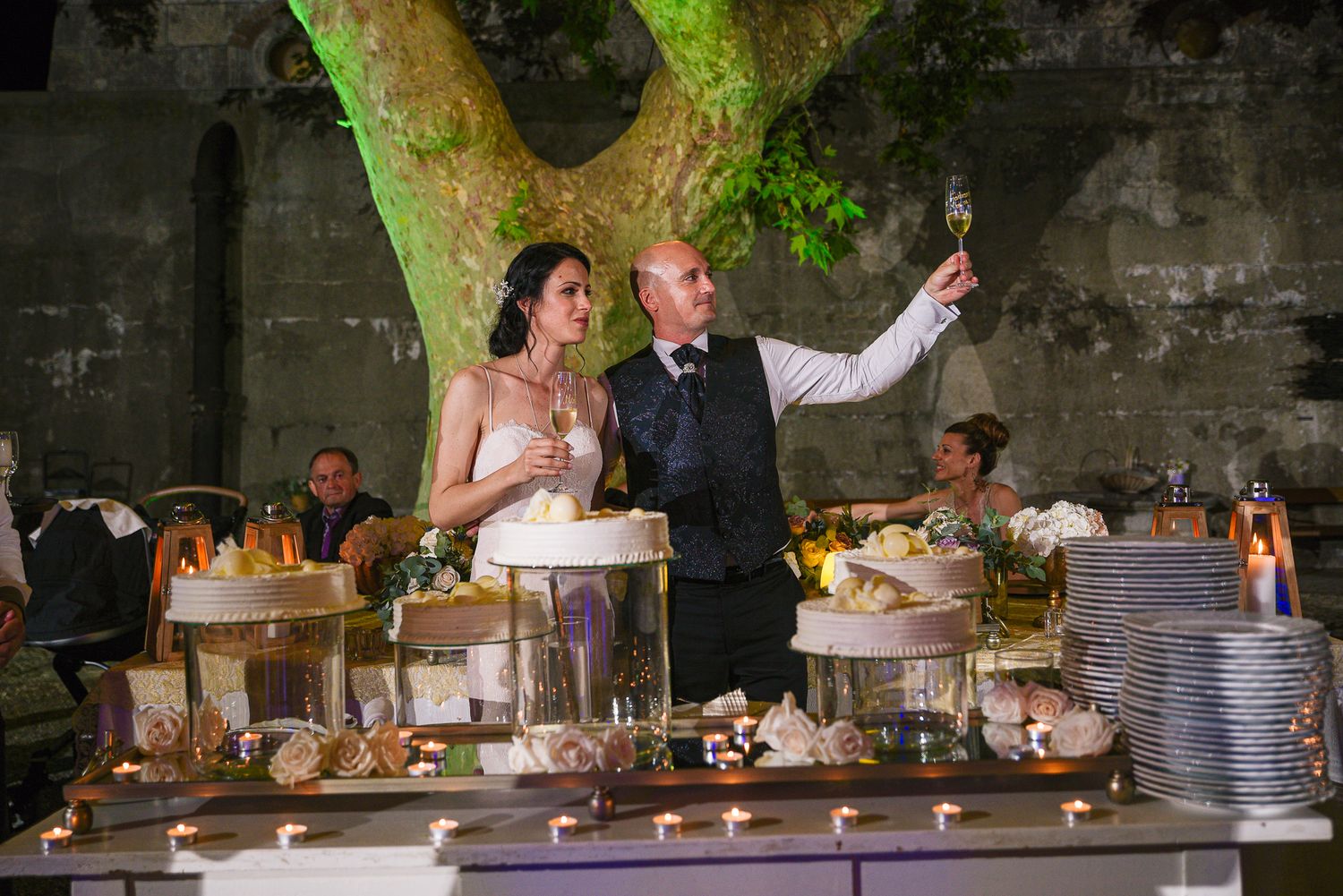 Bride and groom raise champagne glasses beside a decorated wedding cake table under a glowing tree.