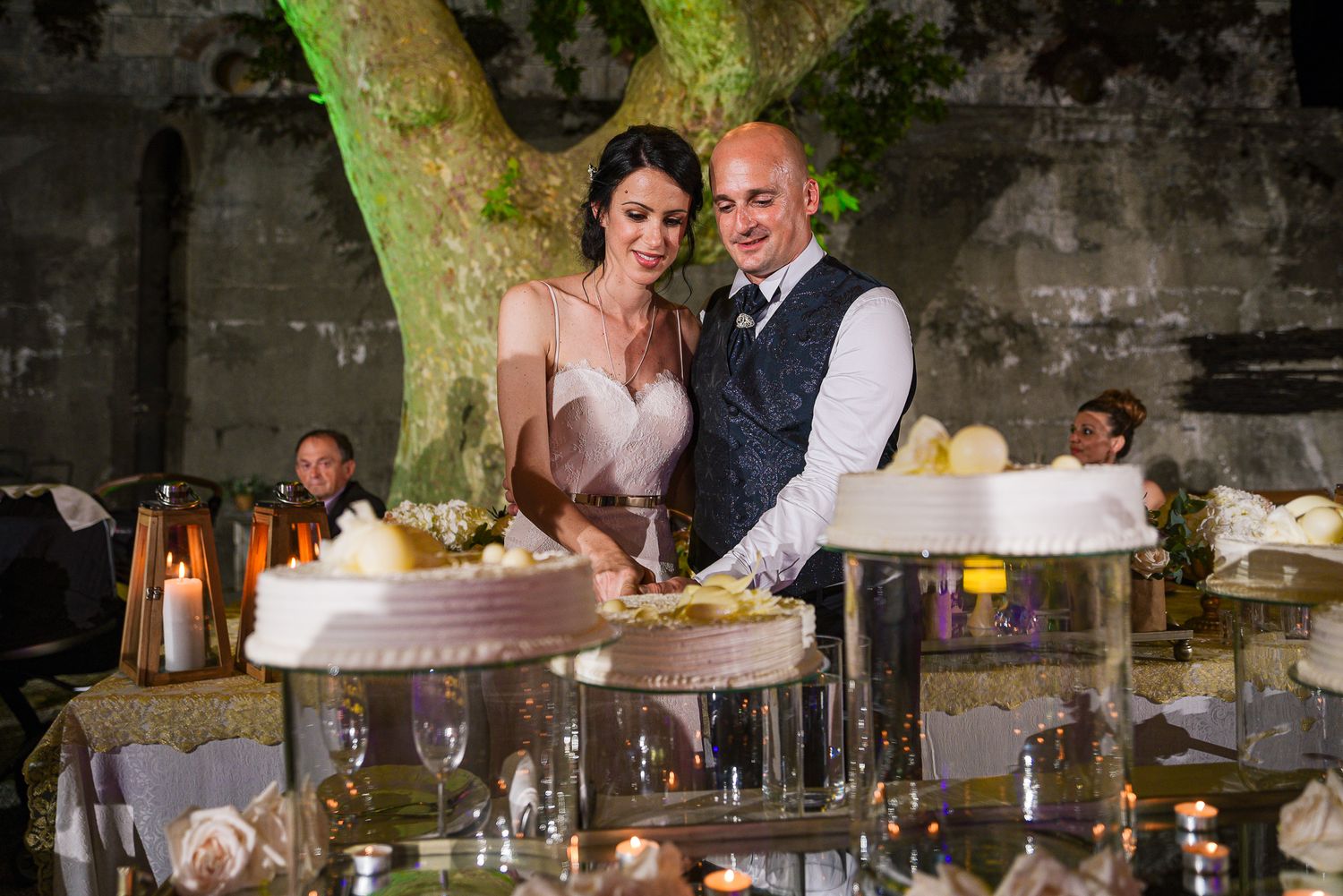 Bride and groom cutting their wedding cake surrounded by candles and floral decorations at night.