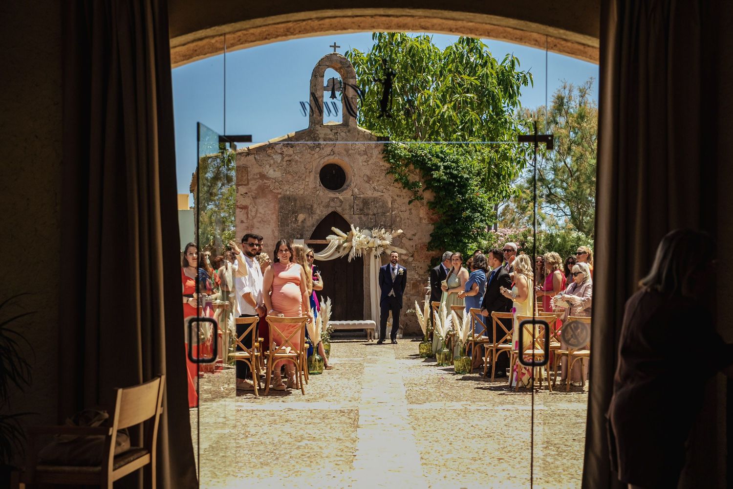 Outdoor wedding ceremony viewed through arched doorway with guests seated along aisle leading to rustic chapel.
