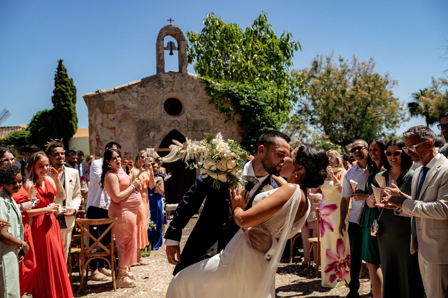 Bride and groom share a romantic kiss at outdoor wedding ceremony with stone chapel and guests applauding.