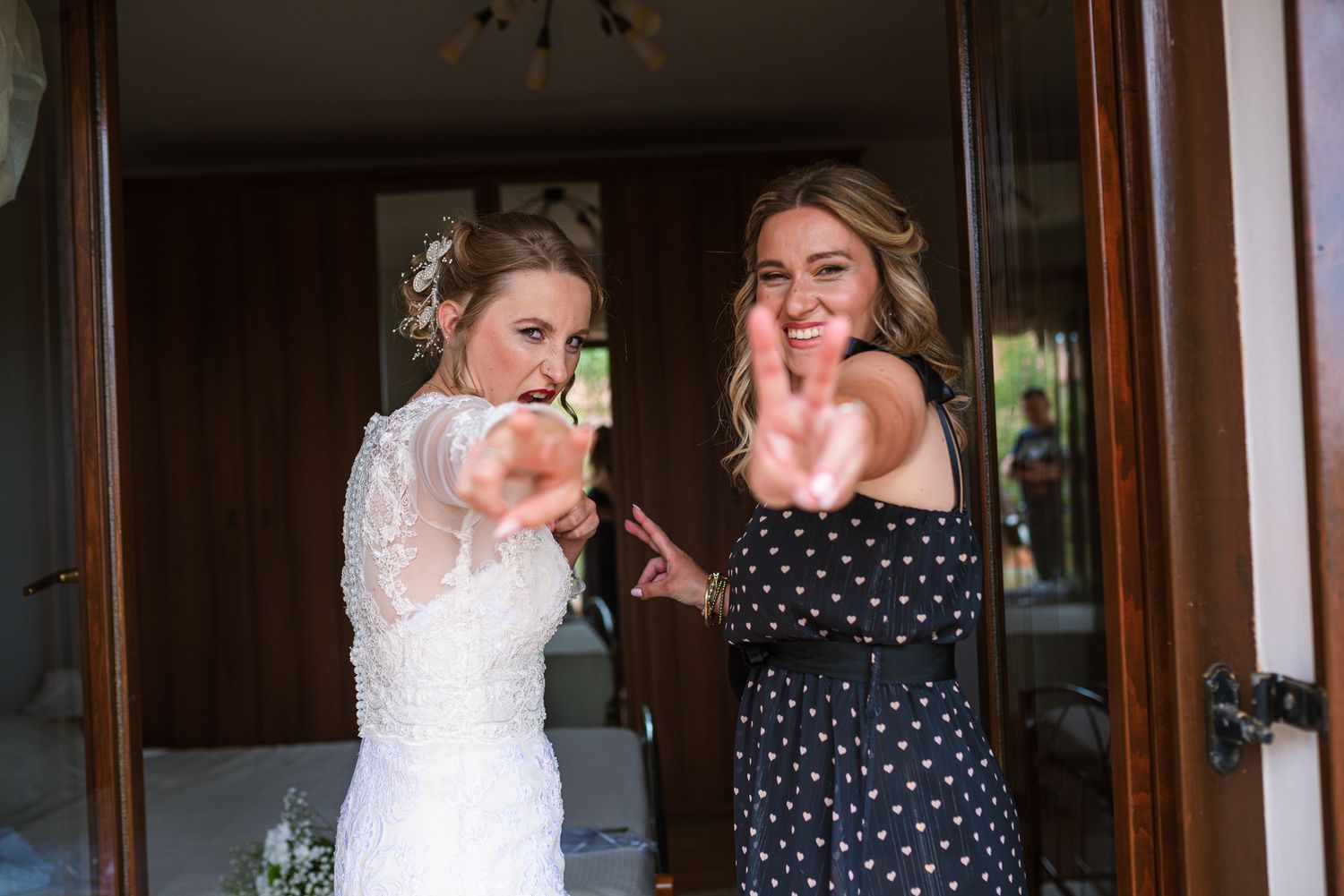 Bride in lace gown and bridesmaid in polka dot dress smiling and making peace signs at wedding celebration.