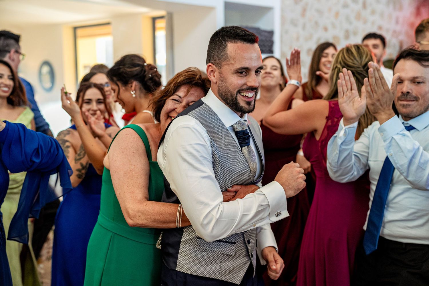 Groom in grey vest celebrates joyfully with wedding guests during a lively reception party.