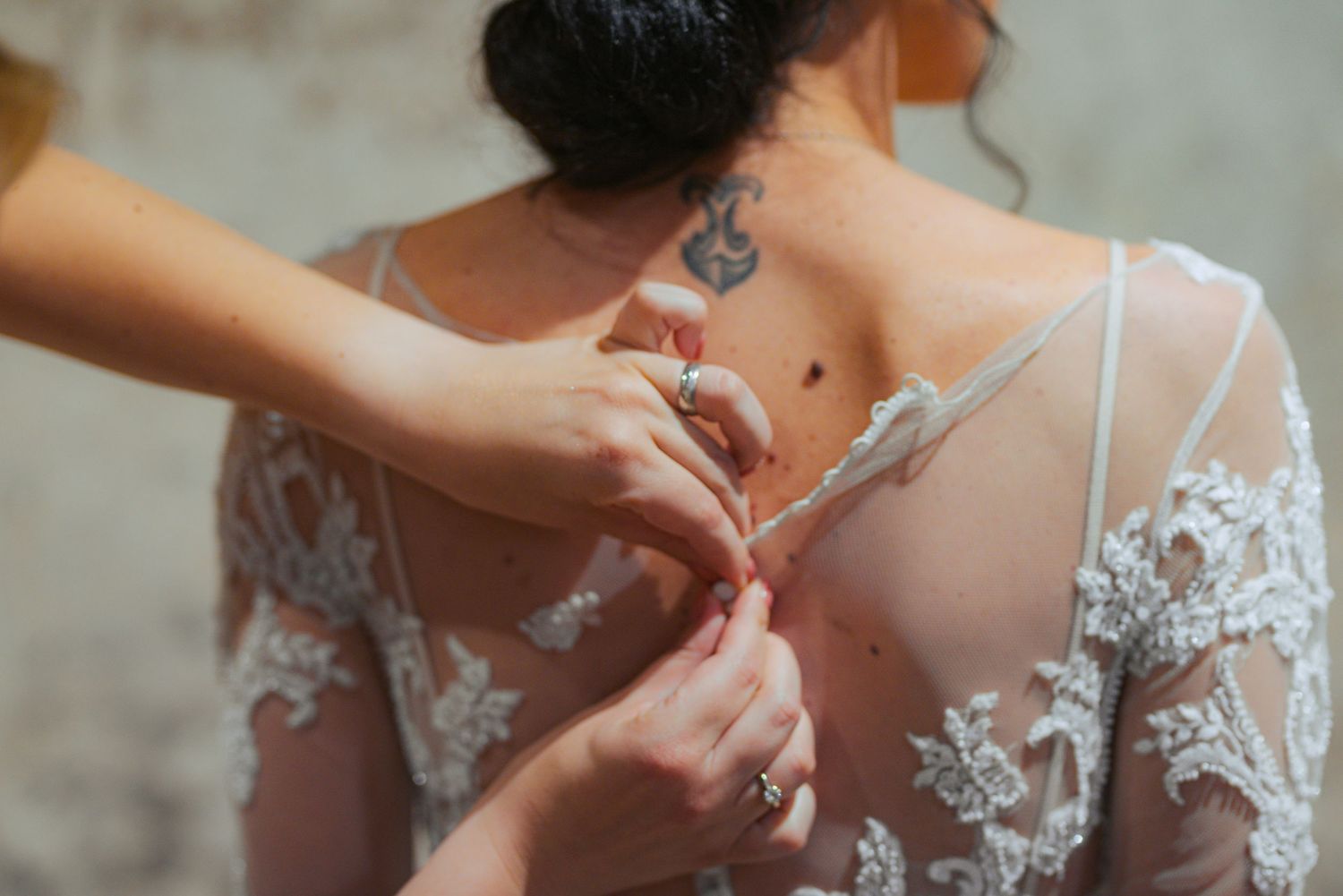 Hands carefully buttoning intricate lace wedding dress on bride with floral back tattoo.