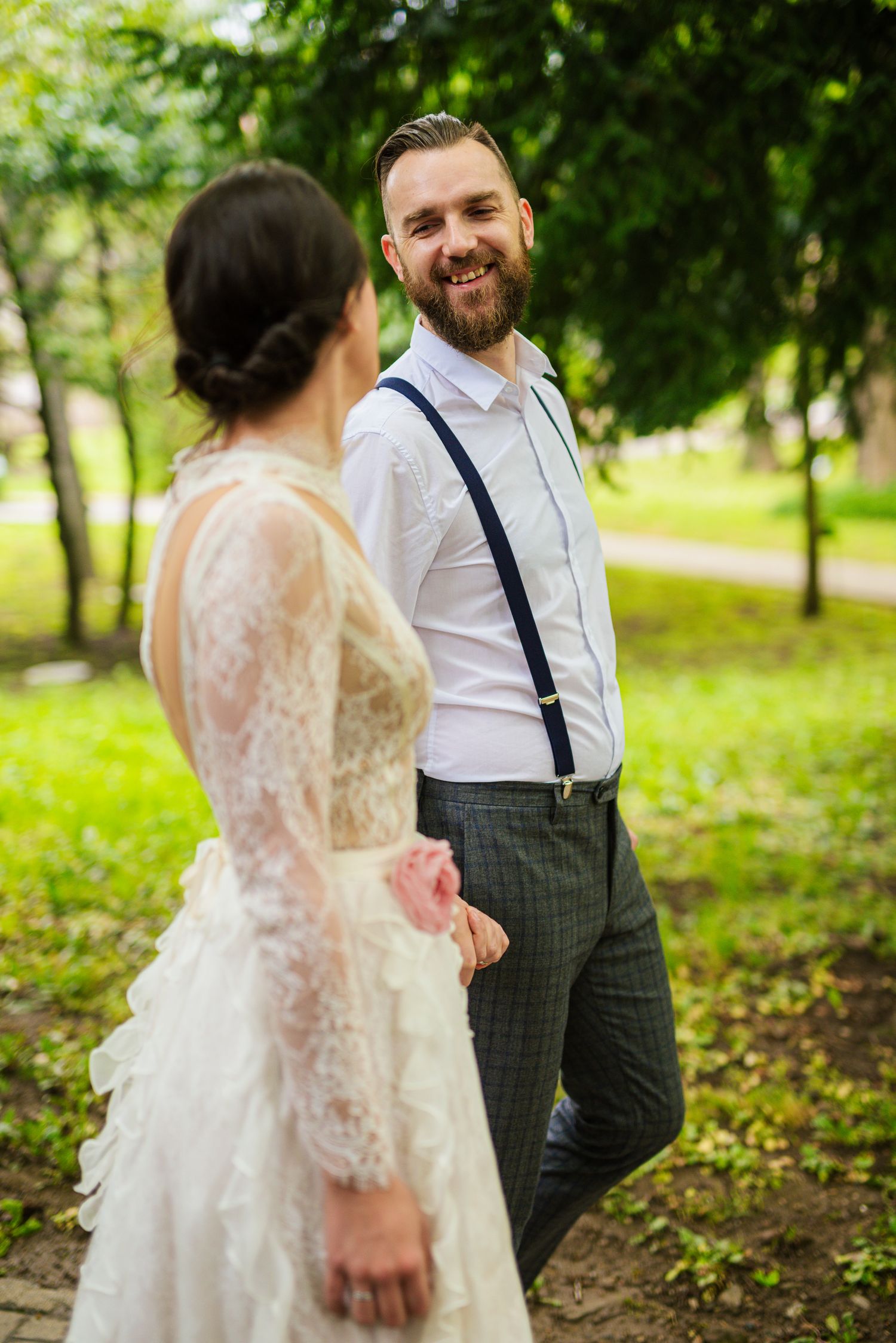 Smiling bearded groom in suspenders holds bride's hand while walking through a lush green park.