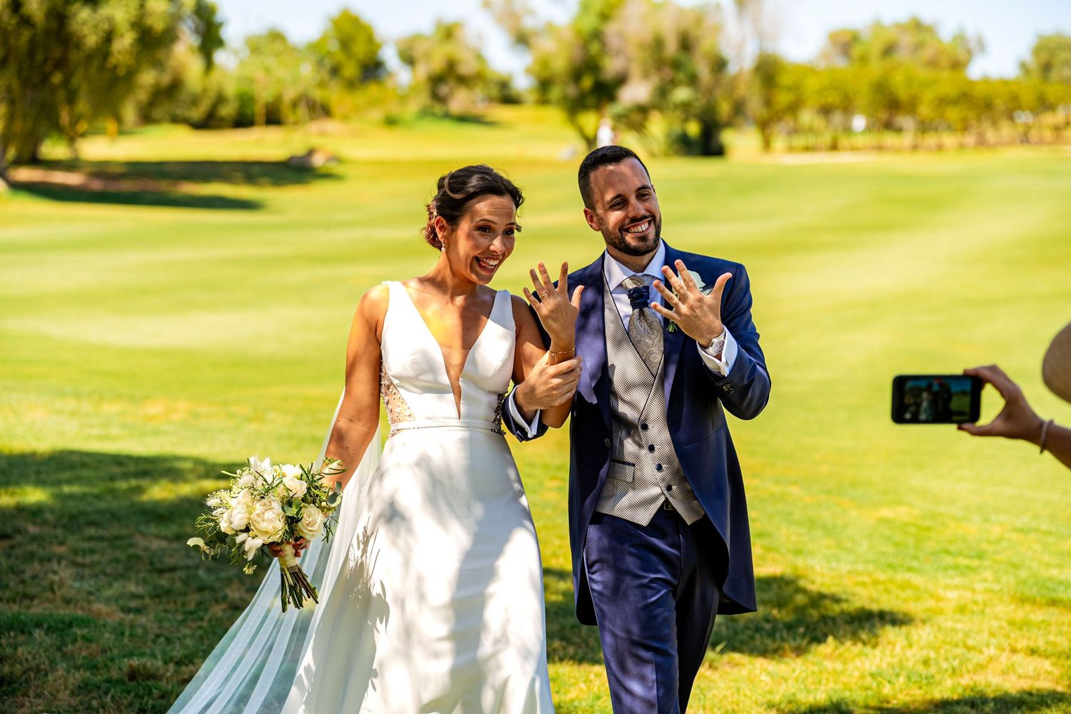 Joyful newlyweds show off wedding rings while walking on golf course as photographer captures moment.