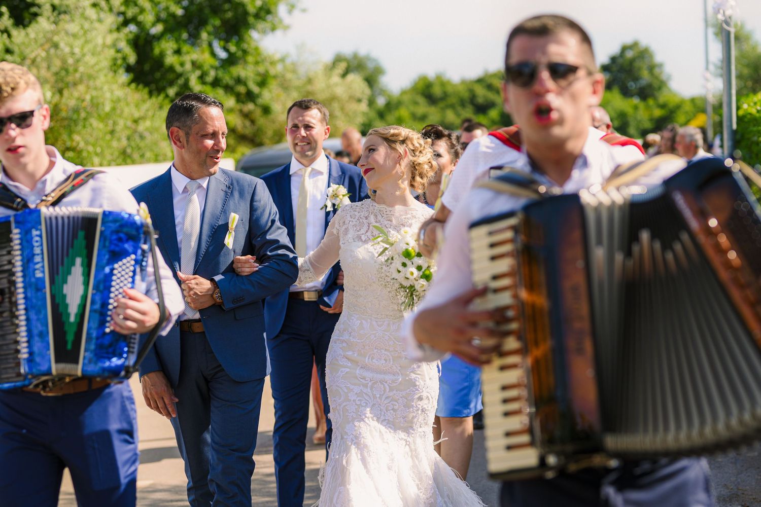 Bride and groom walking outdoors accompanied by accordion musicians during traditional wedding procession.