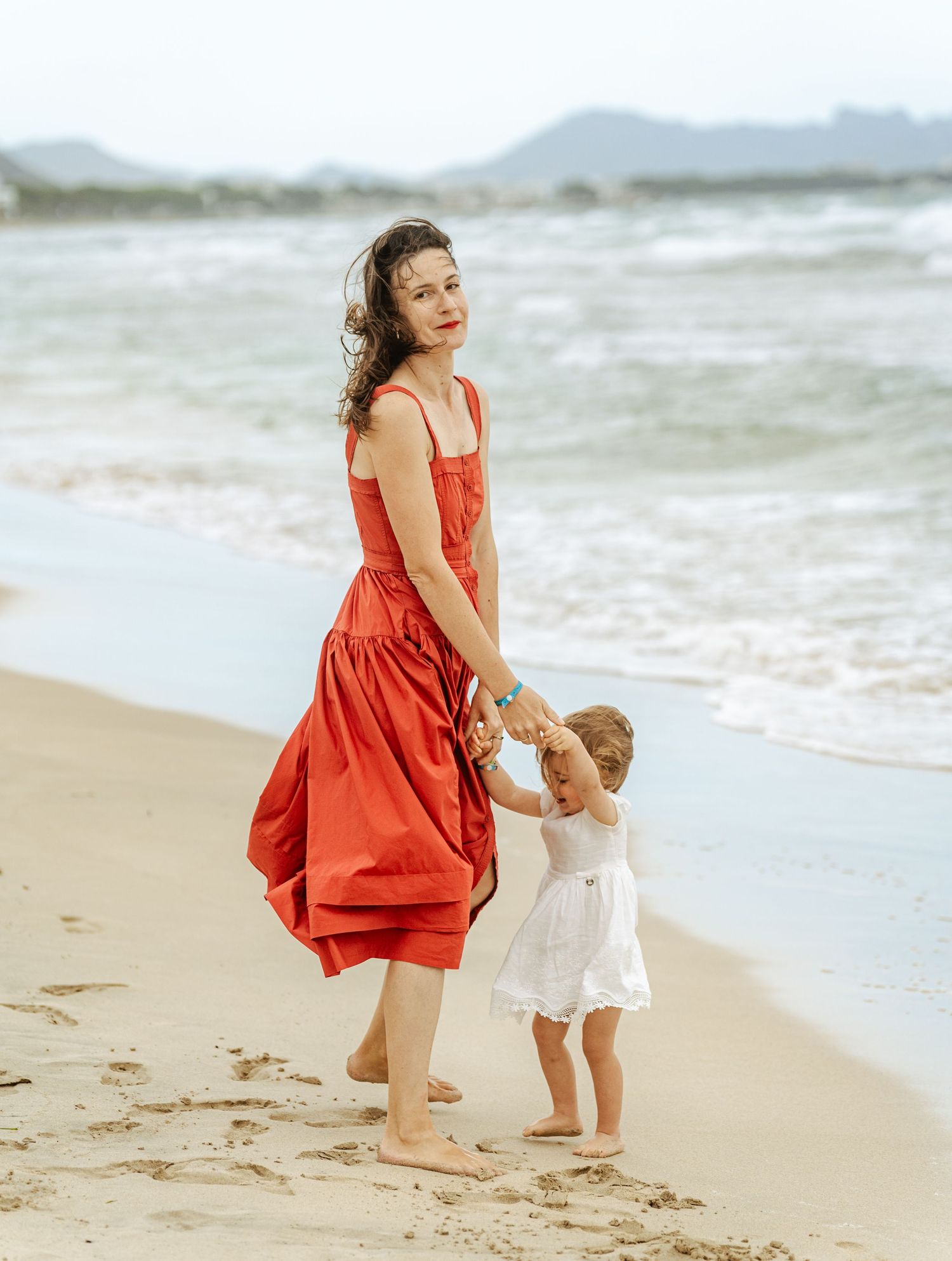 Mother in red dress holding young daughter's hand while walking on sandy beach.