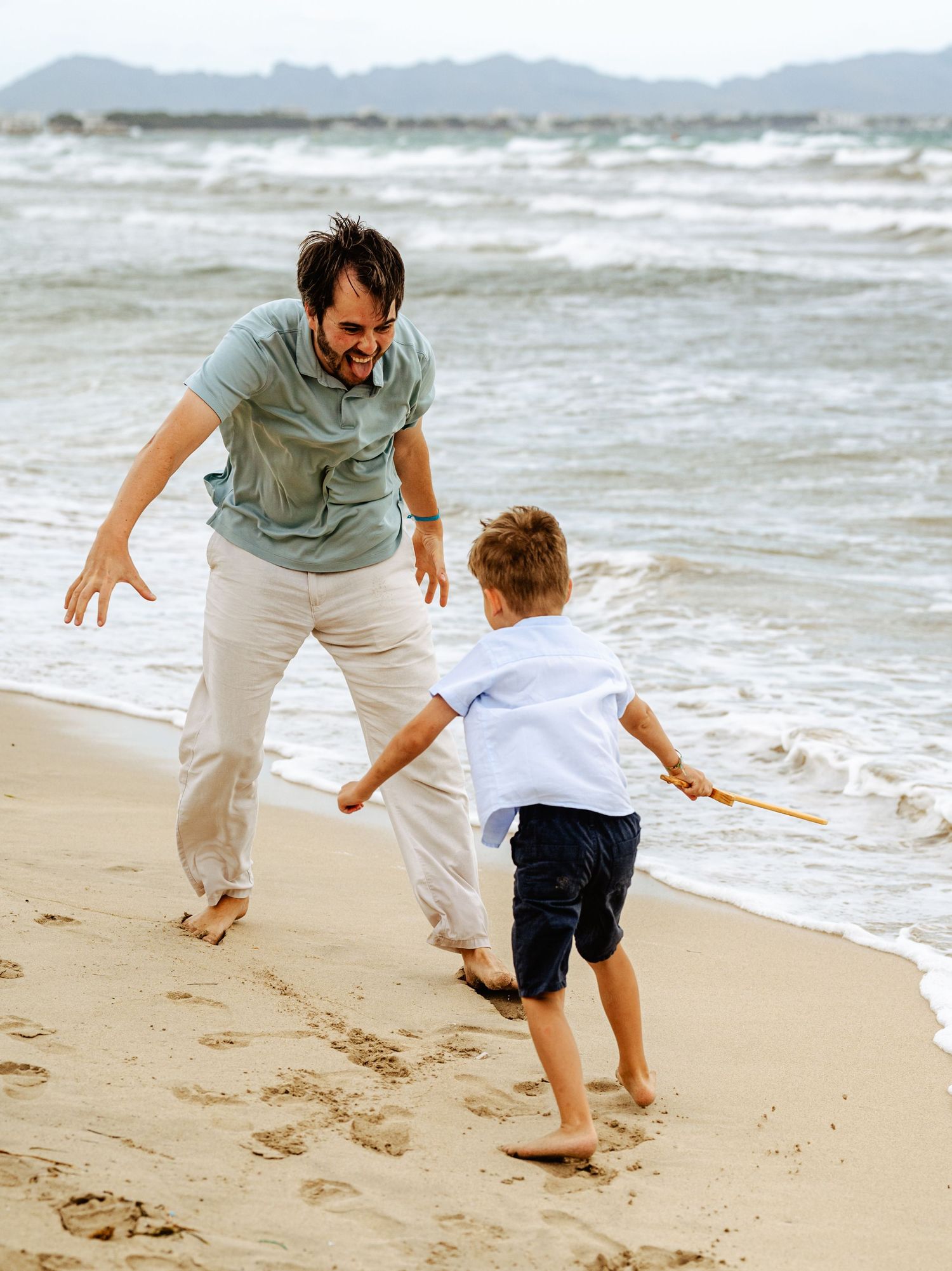 Father and son playing chase on sandy beach with ocean waves in background.