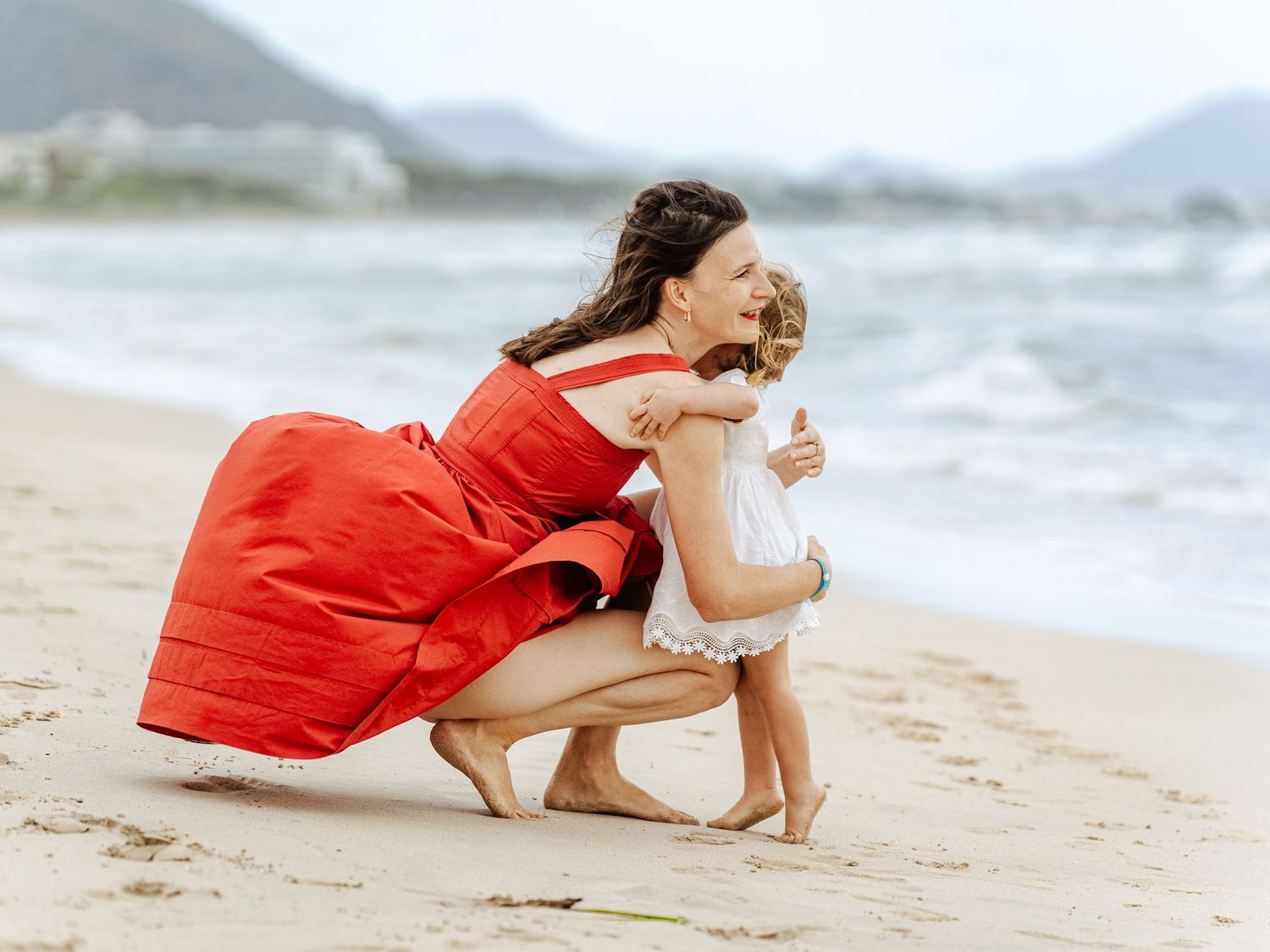 Mother in red dress hugs toddler girl on sandy beach with mountains.