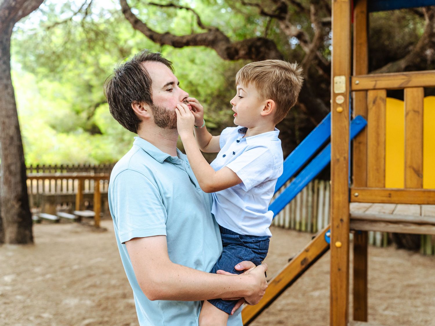Father holding his young son at a playground, the boy touching his dad's face playfully.
