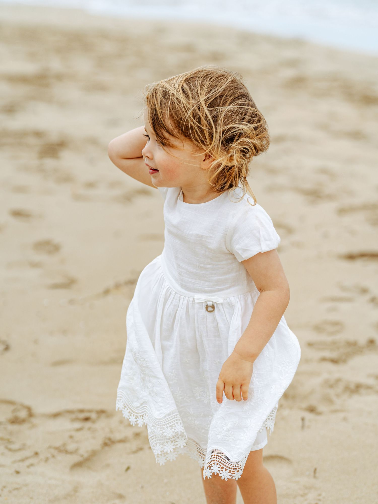 A toddler girl in a white lace dress stands on a sandy beach with her hand in her wind-blown hair.