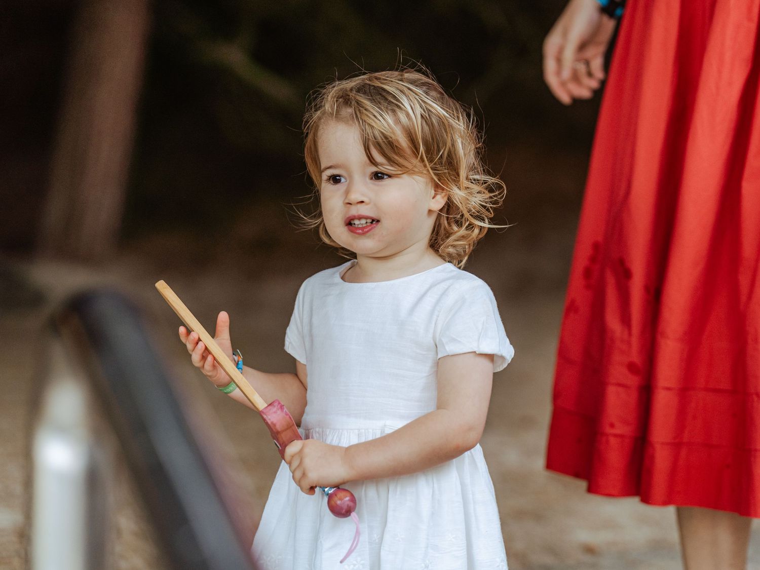 Smiling toddler girl in white dress holding a colorful wooden toy outdoors near a red skirt.