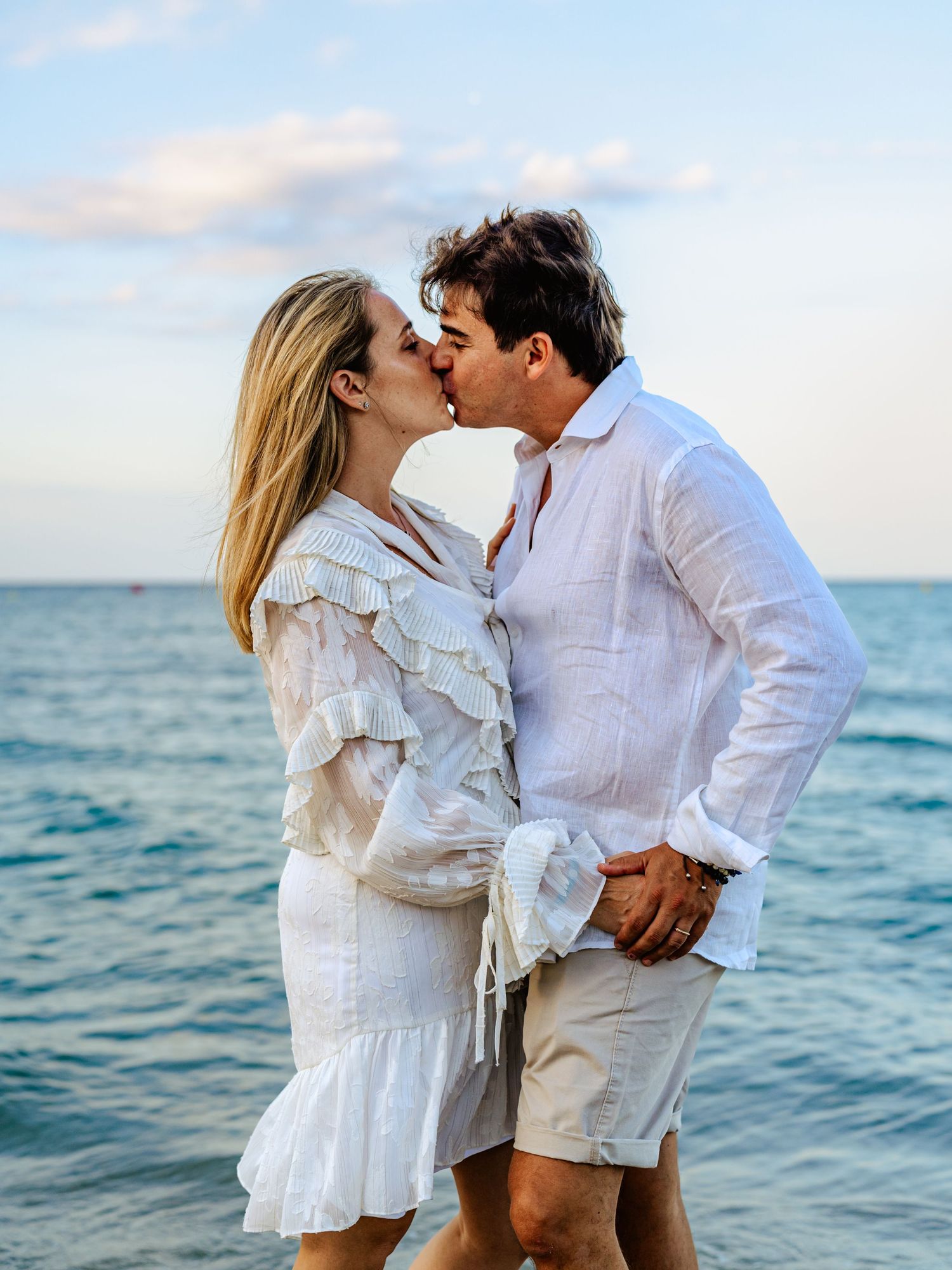 Couple kissing on sunny beach wearing matching white outfits with ocean in background.
