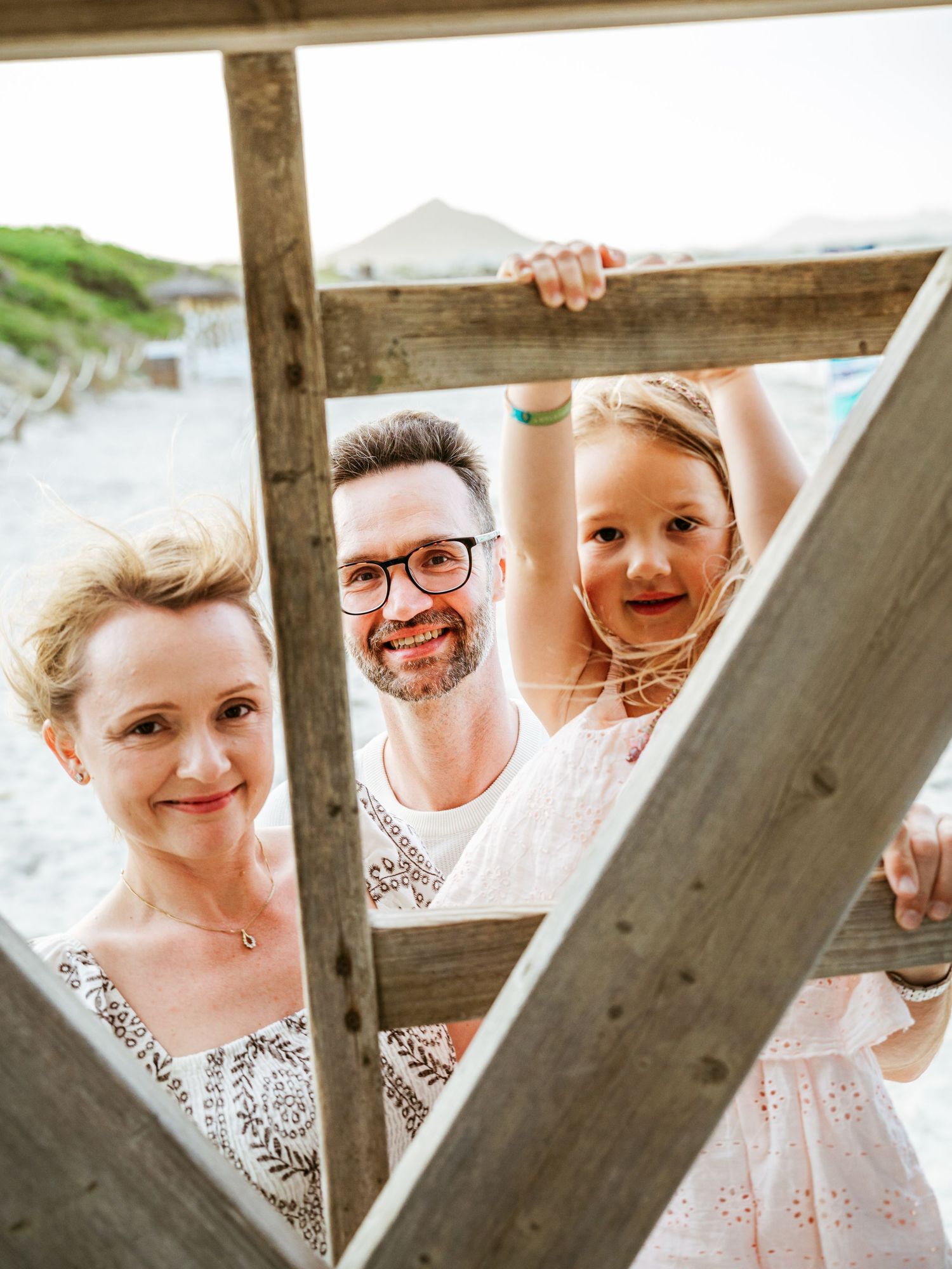 Cheerful family of three peering through wooden beach structure frames, smiling at camera.