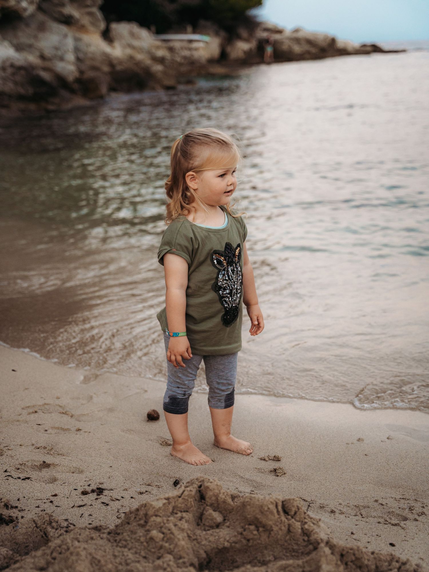 Cute blonde toddler girl in olive green shirt standing barefoot on sandy beach by calm water.