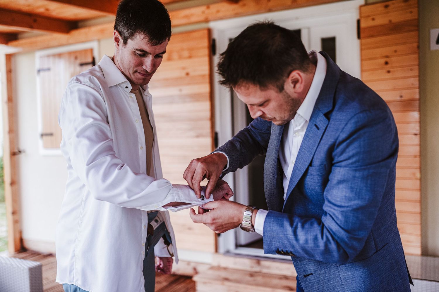 Groom in blue suit having cufflinks fastened by groomsman outside wooden building.
