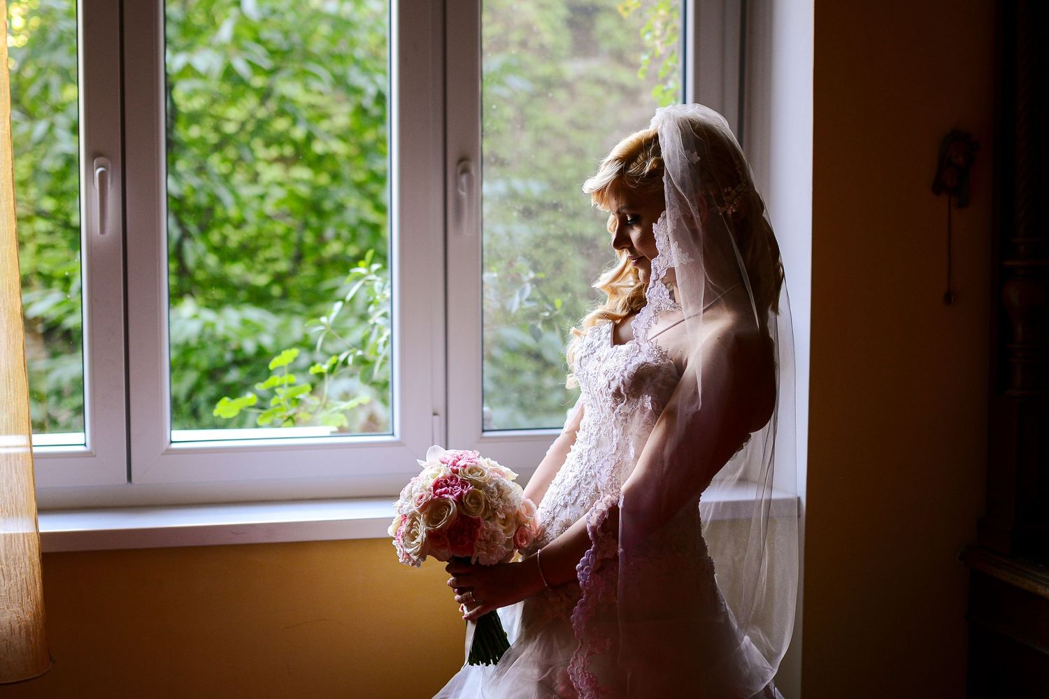 Bride in a lace gown and veil gazes out a window while holding a pink bouquet.