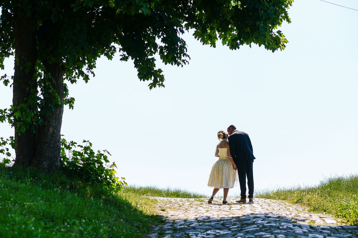 Bride and groom walking hand in hand on a cobblestone path beneath a large green tree.