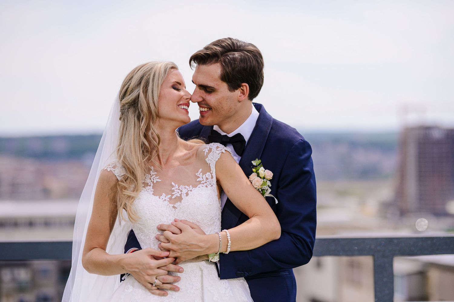 Happy newlyweds in lace wedding dress and blue suit sharing a romantic rooftop moment.